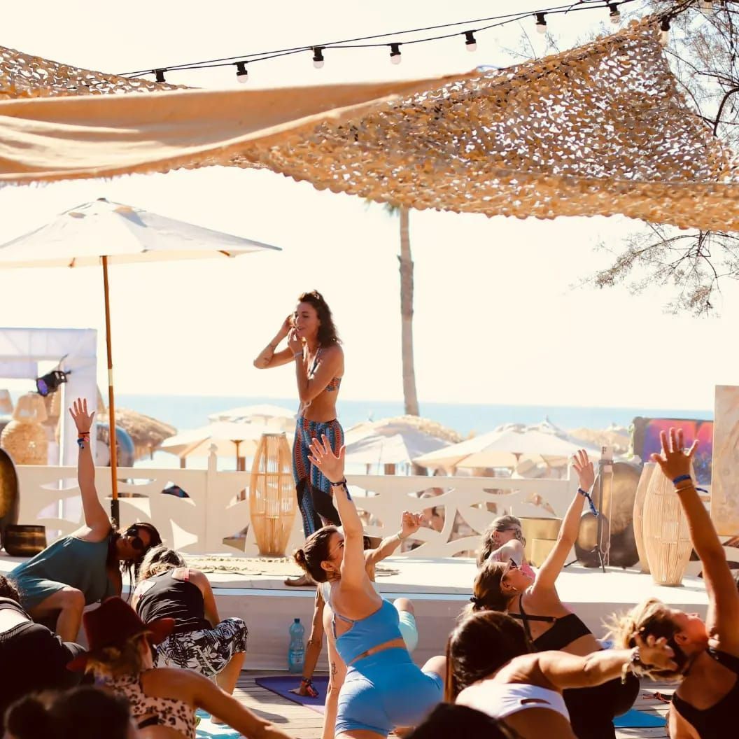 People doing yoga on a beach, led by an instructor. Bright sunny day, open air space.