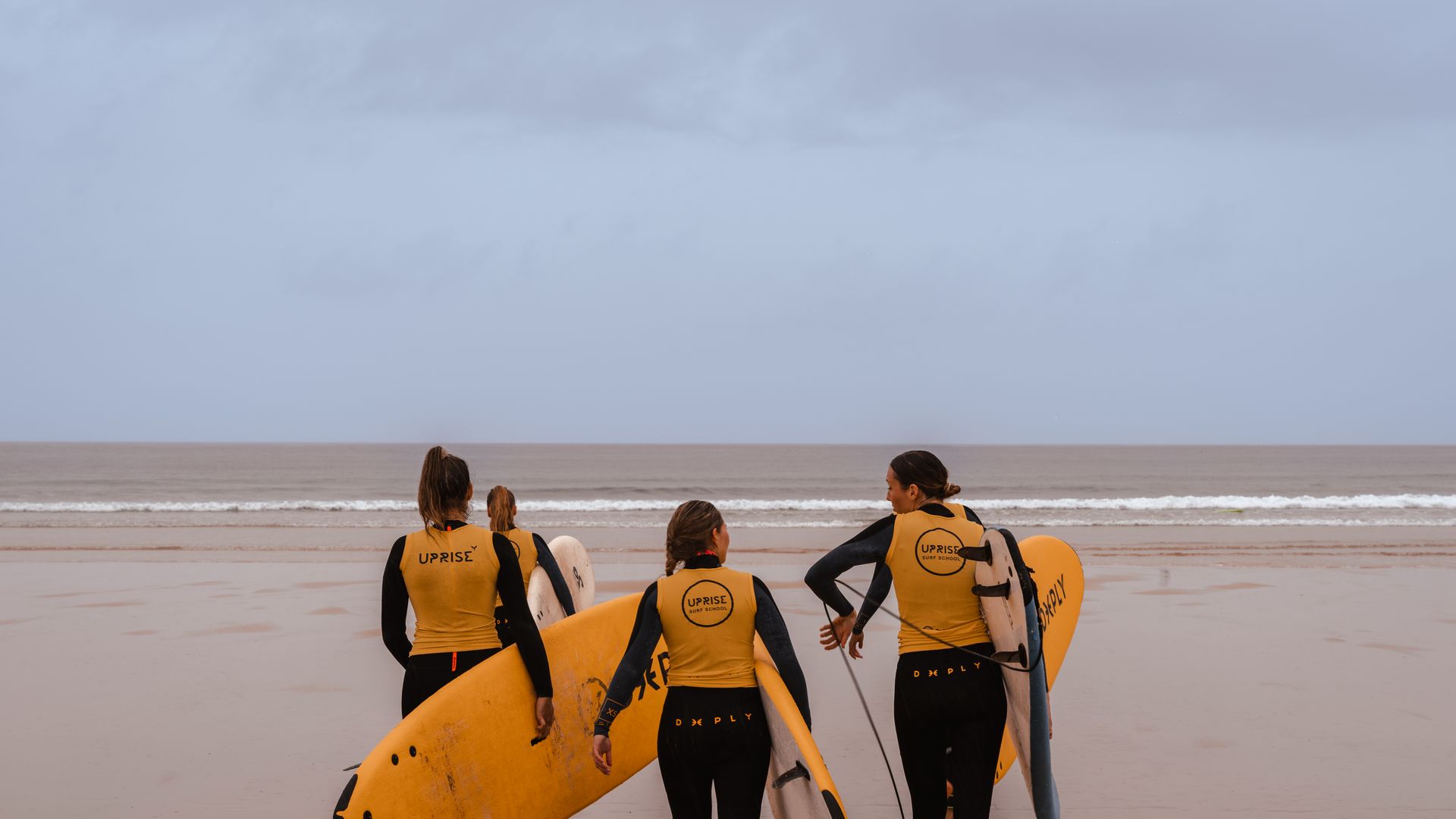 Surfers with yellow vests carry surfboards on a sandy beach under an overcast sky.