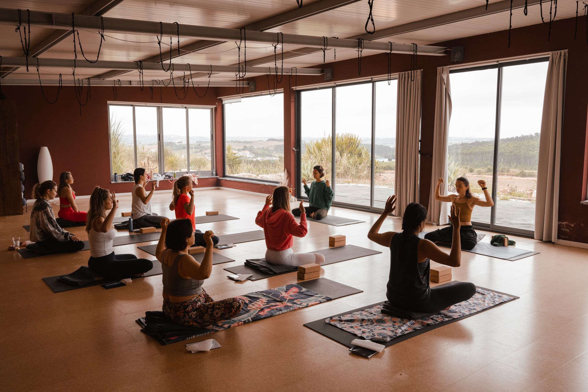 Yoga class in a studio with large windows overlooking a landscape. People seated on mats, some with hands raised, led by an instructor.