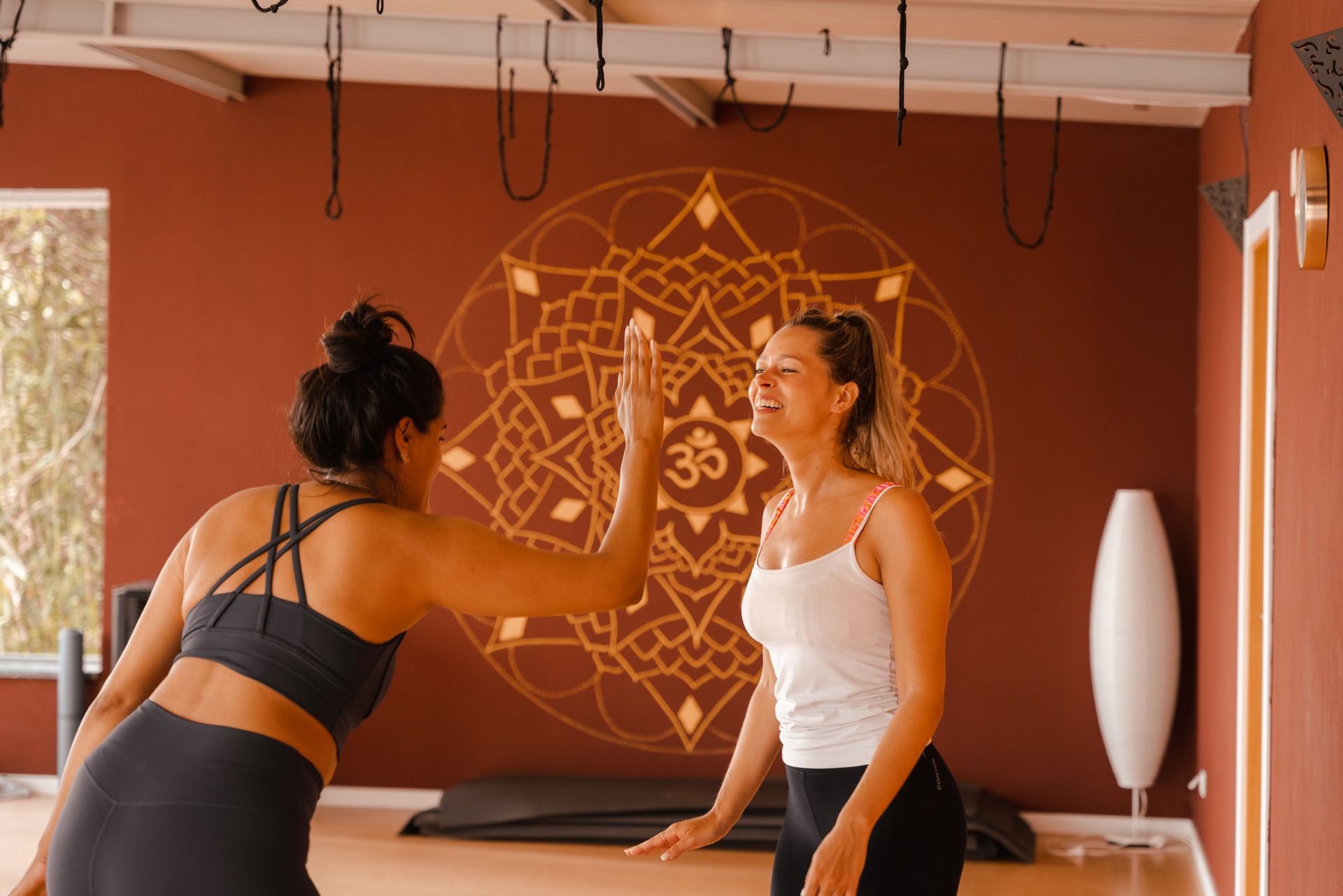Two women high-fiving in a yoga studio with a large mandala design on the wall.