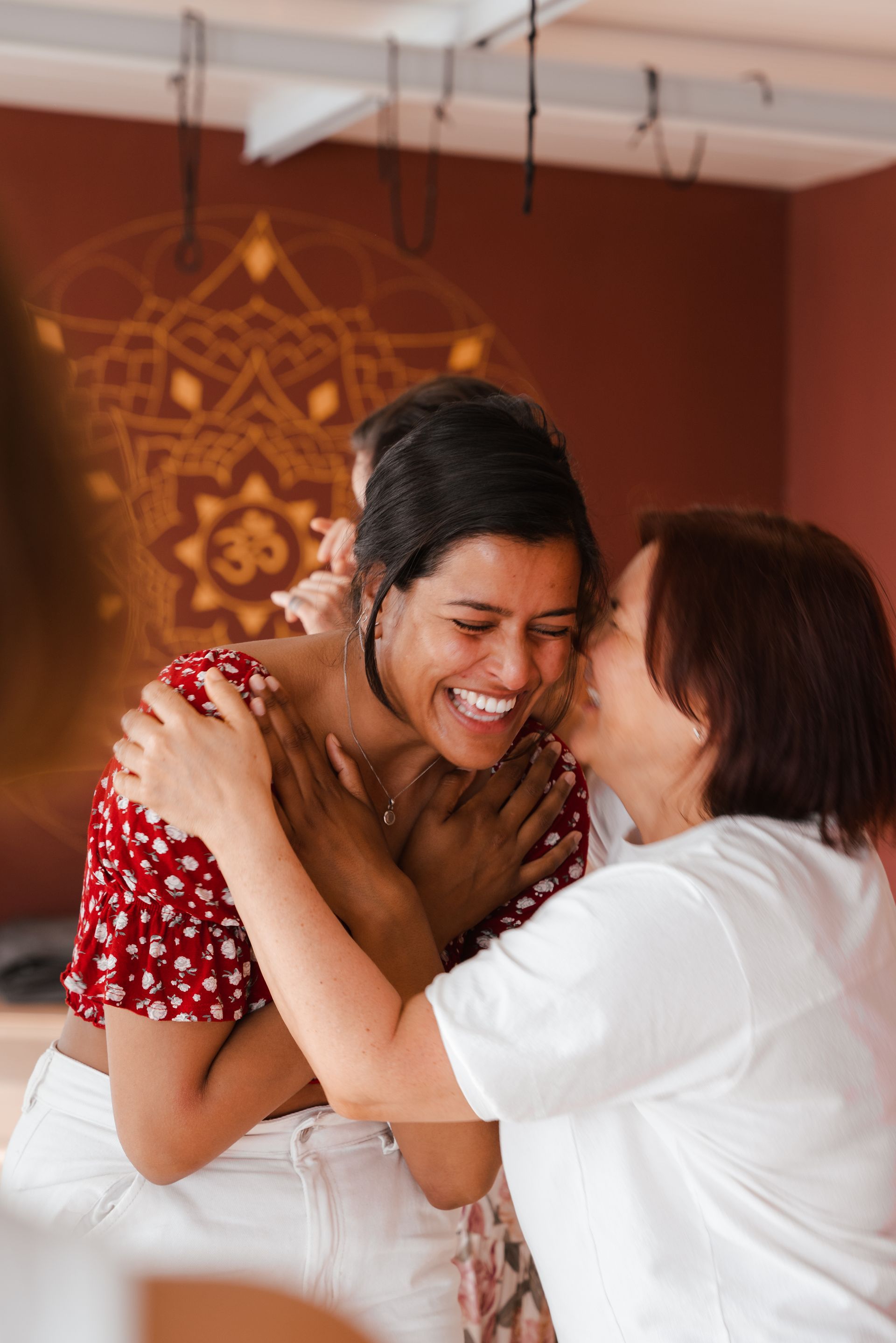Woman hugs and whispers to another woman laughing, inside a room with mandala art.