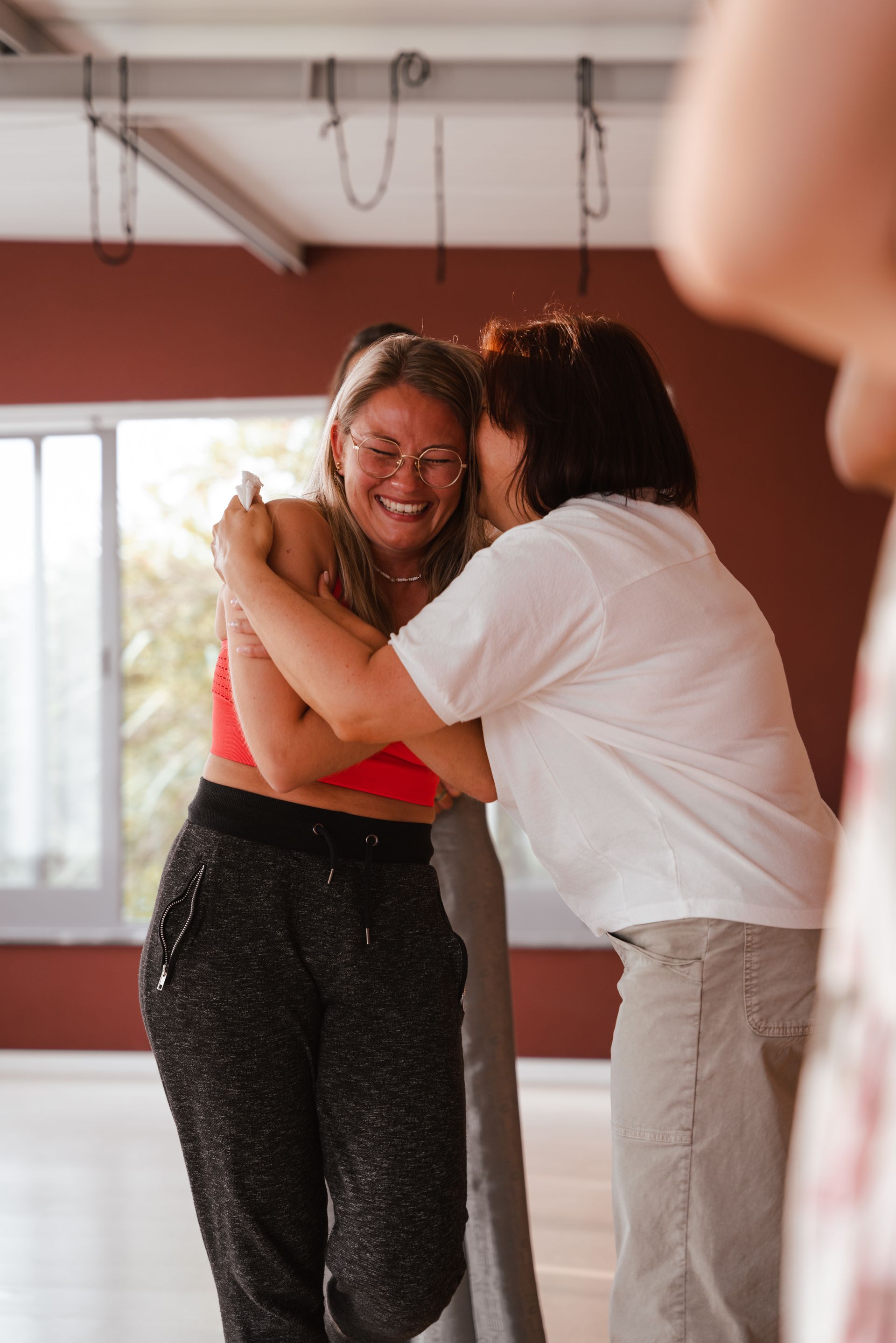 Two women embracing indoors; one laughs, wearing a red top and black pants.