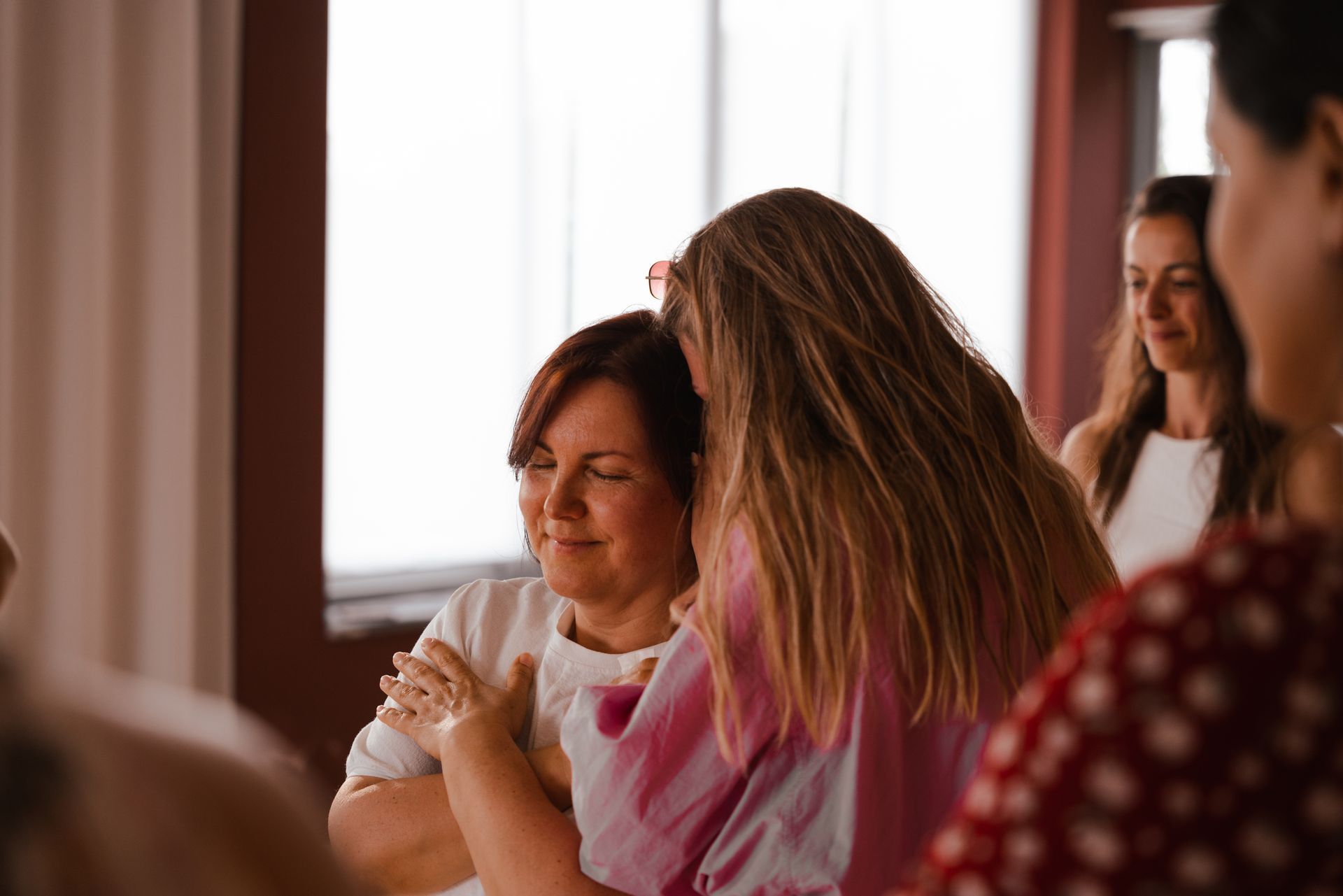 Woman with closed eyes is embraced by another woman, surrounded by others in a bright room.