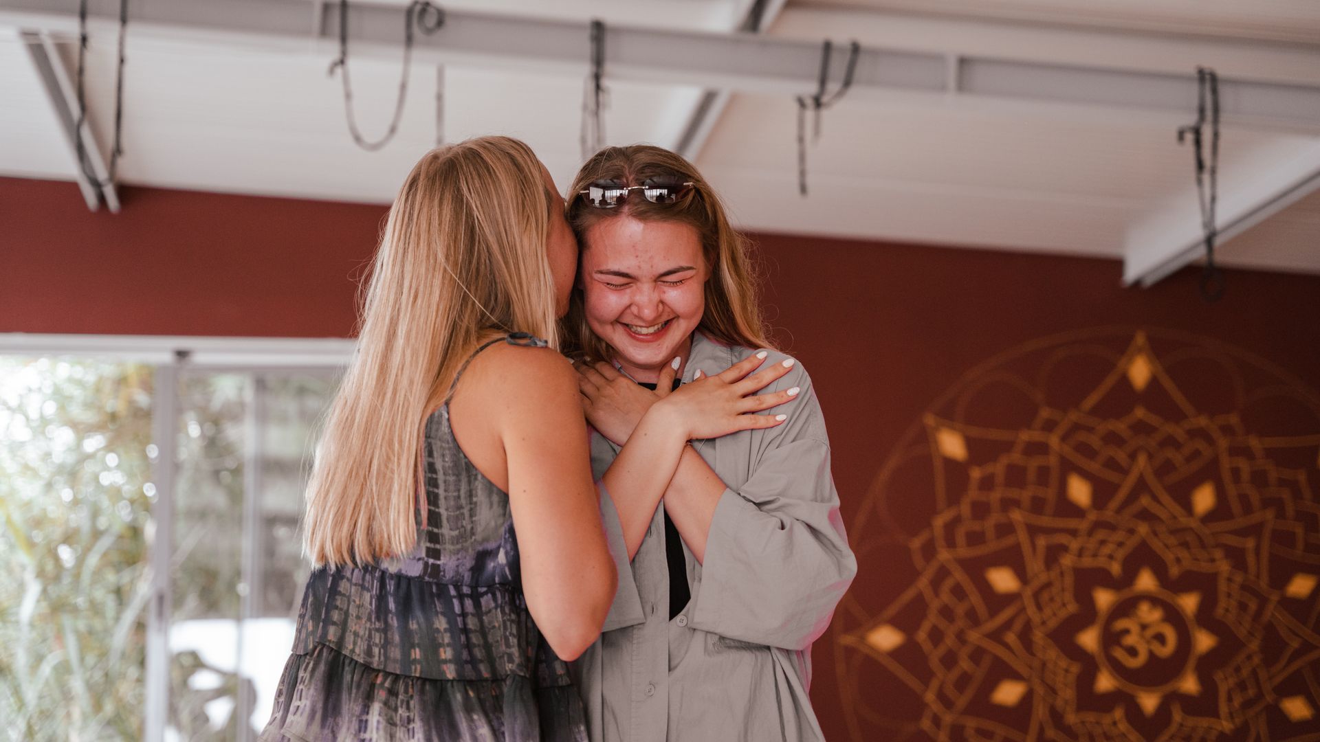 Two women embrace, one whispering in the other's ear, smiling. Indoors, mandala art on wall.