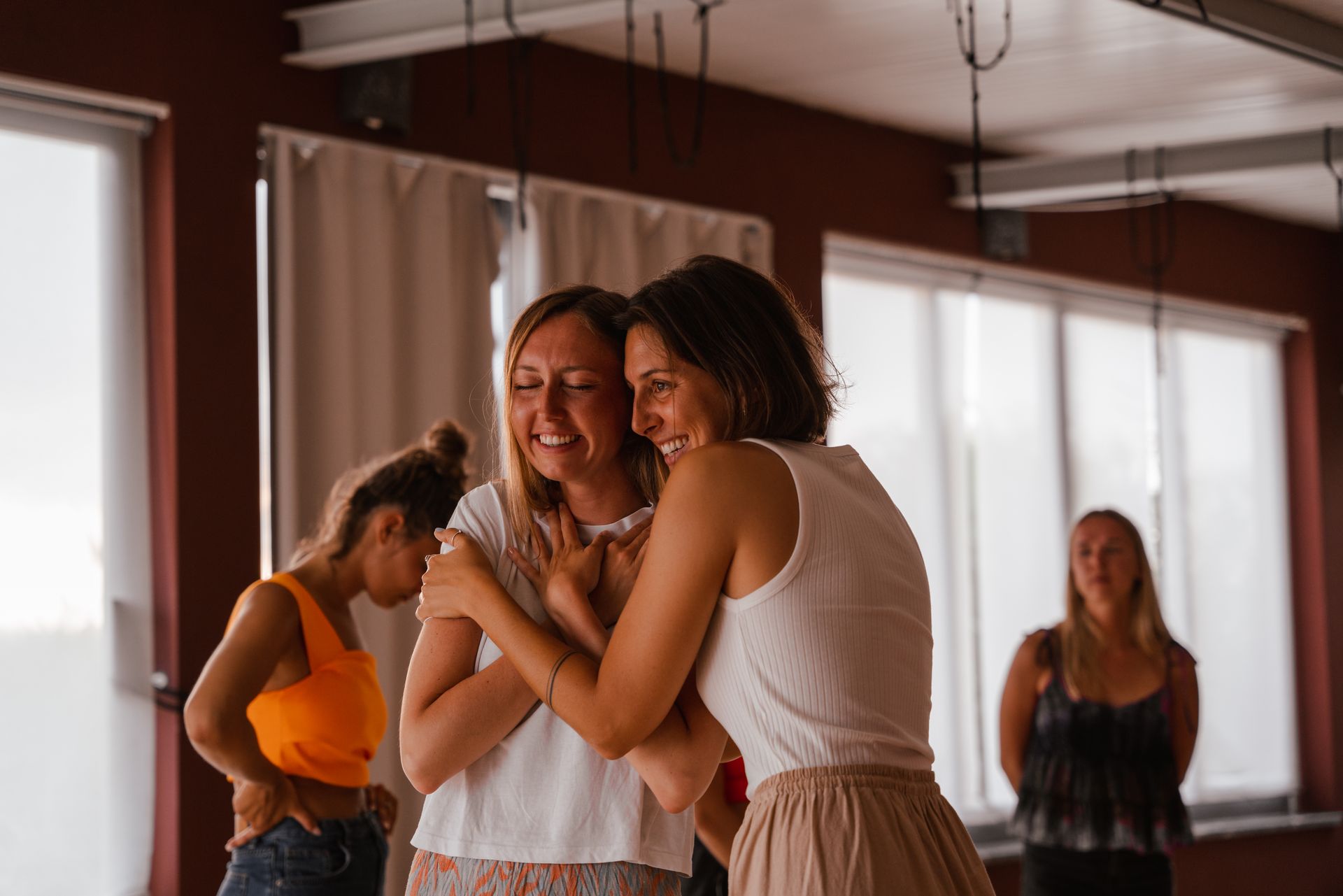 Two women embrace, smiling in a dance studio. Others watch in the background. Natural light.