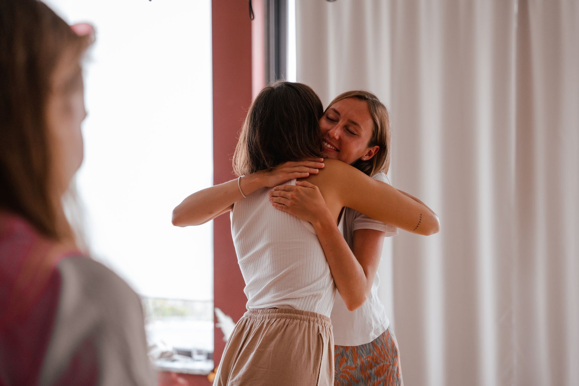 Two women hugging, one smiling. Another woman is partially visible in the background. Bright, indoor setting.