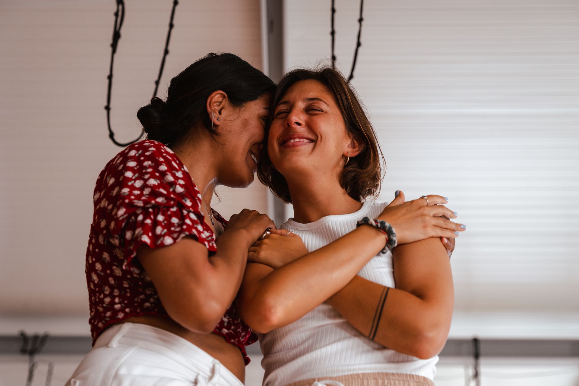 Two people embracing, one whispering in the other's ear, both smiling, with a simple white background.