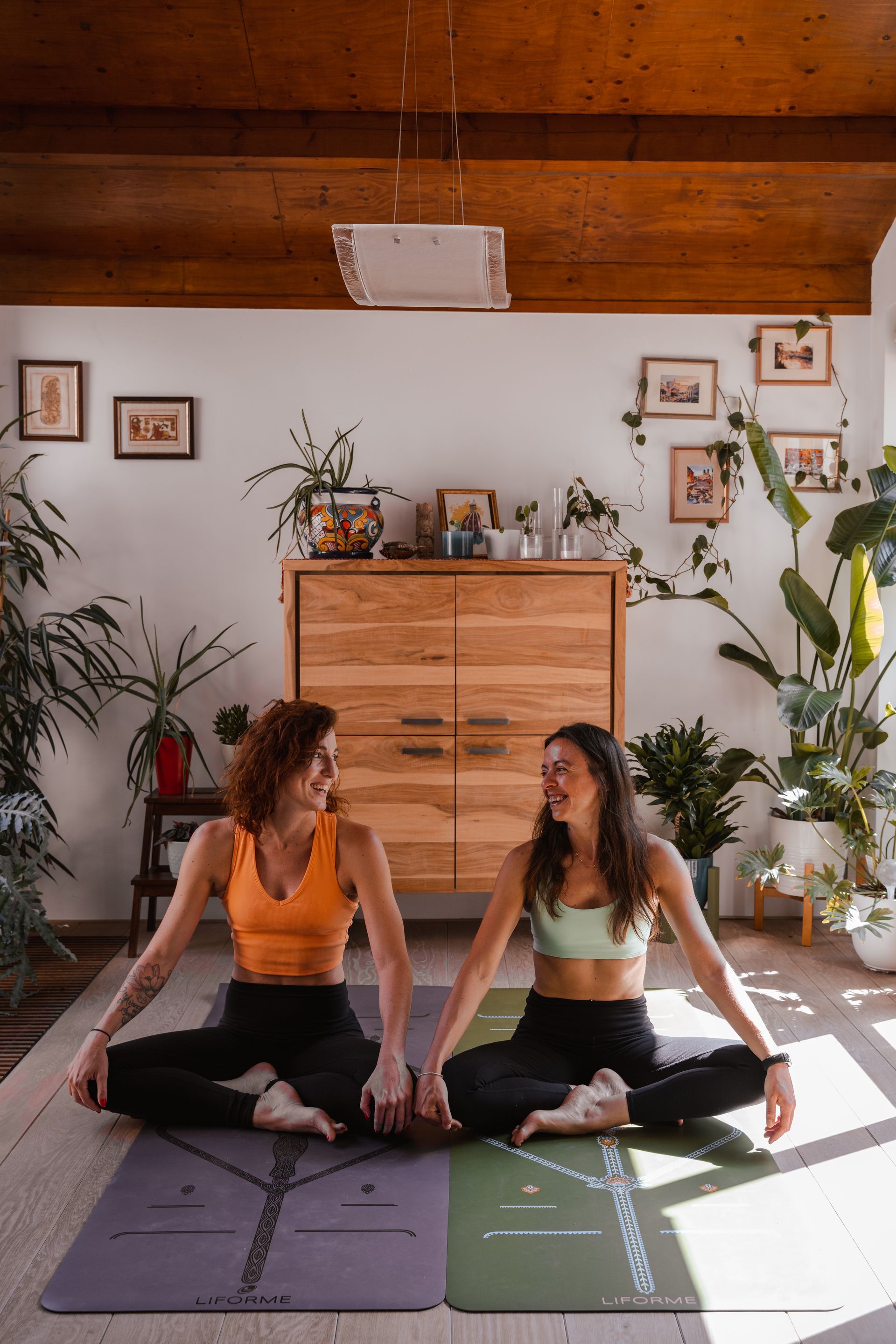 Two women in workout clothes seated on yoga mats, holding hands, smiling. Interior with plants and wooden furniture.