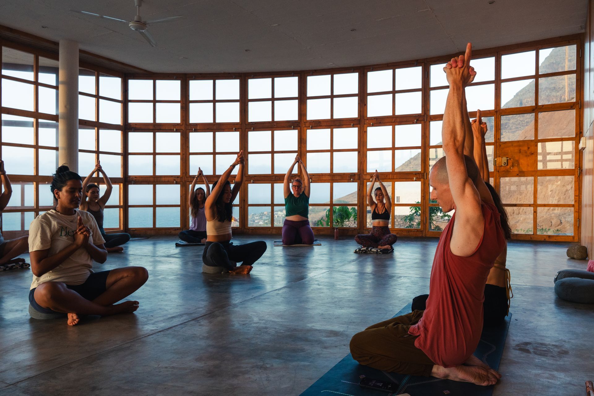 People in a sunlit room doing yoga, raising arms. Large windows, natural light.