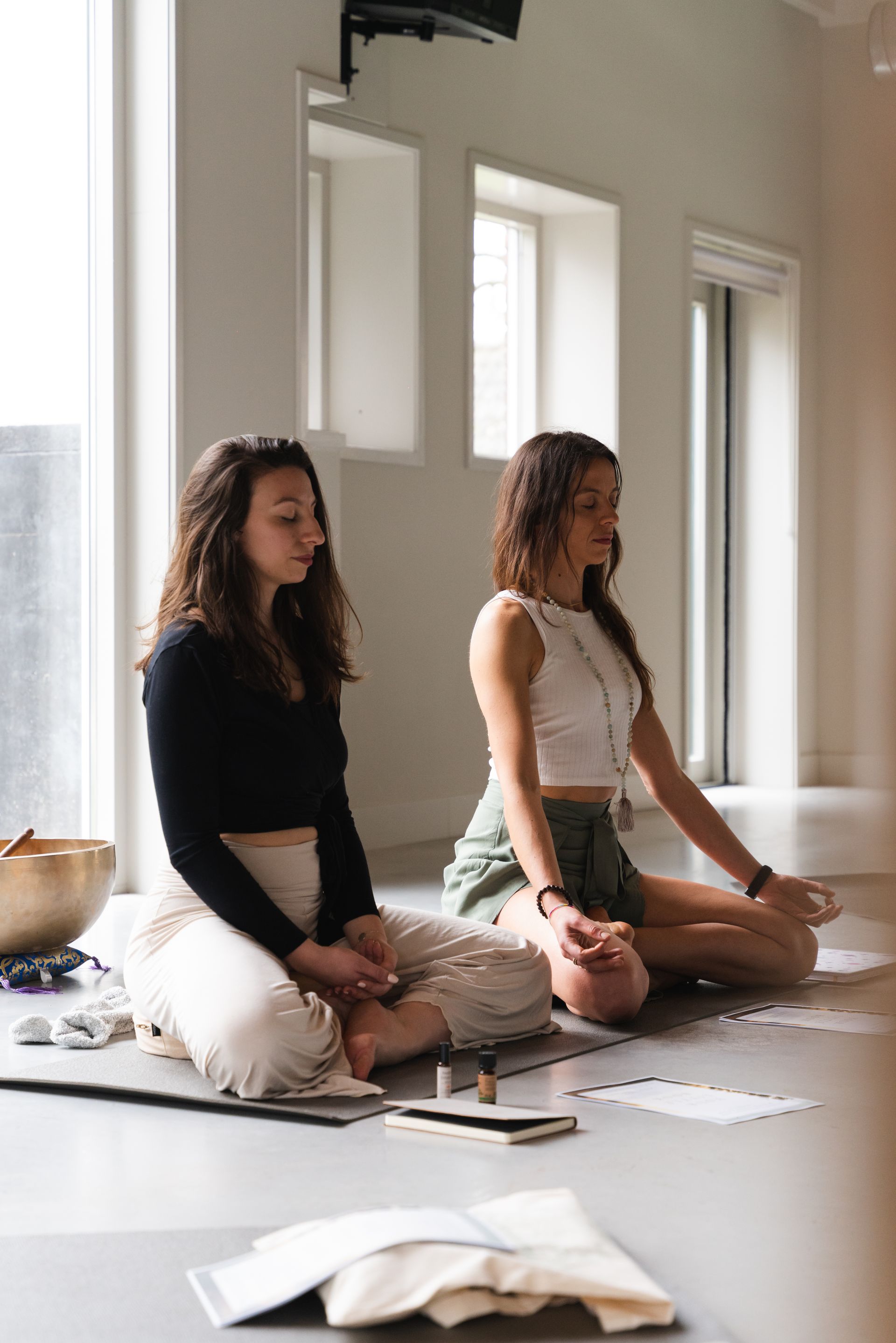Two women meditate in a brightly lit room, eyes closed, hands in lap.