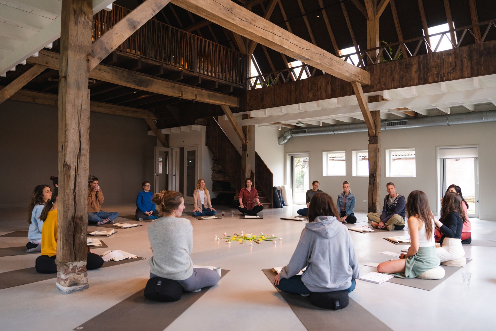 People seated in a circle on mats inside a wooden-beamed building, some meditating, others taking notes.