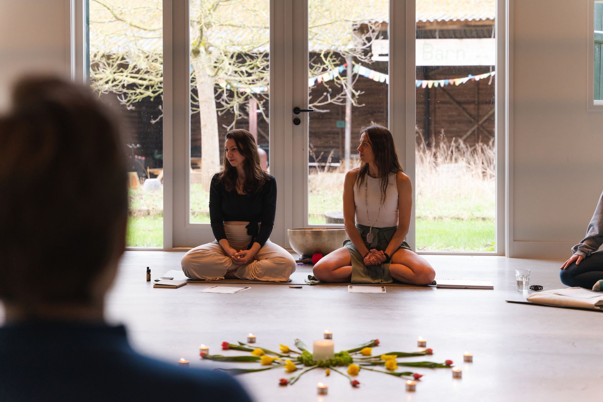 Two women seated on cushions, smiling, with a candle mandala on the floor. In a room with glass doors.