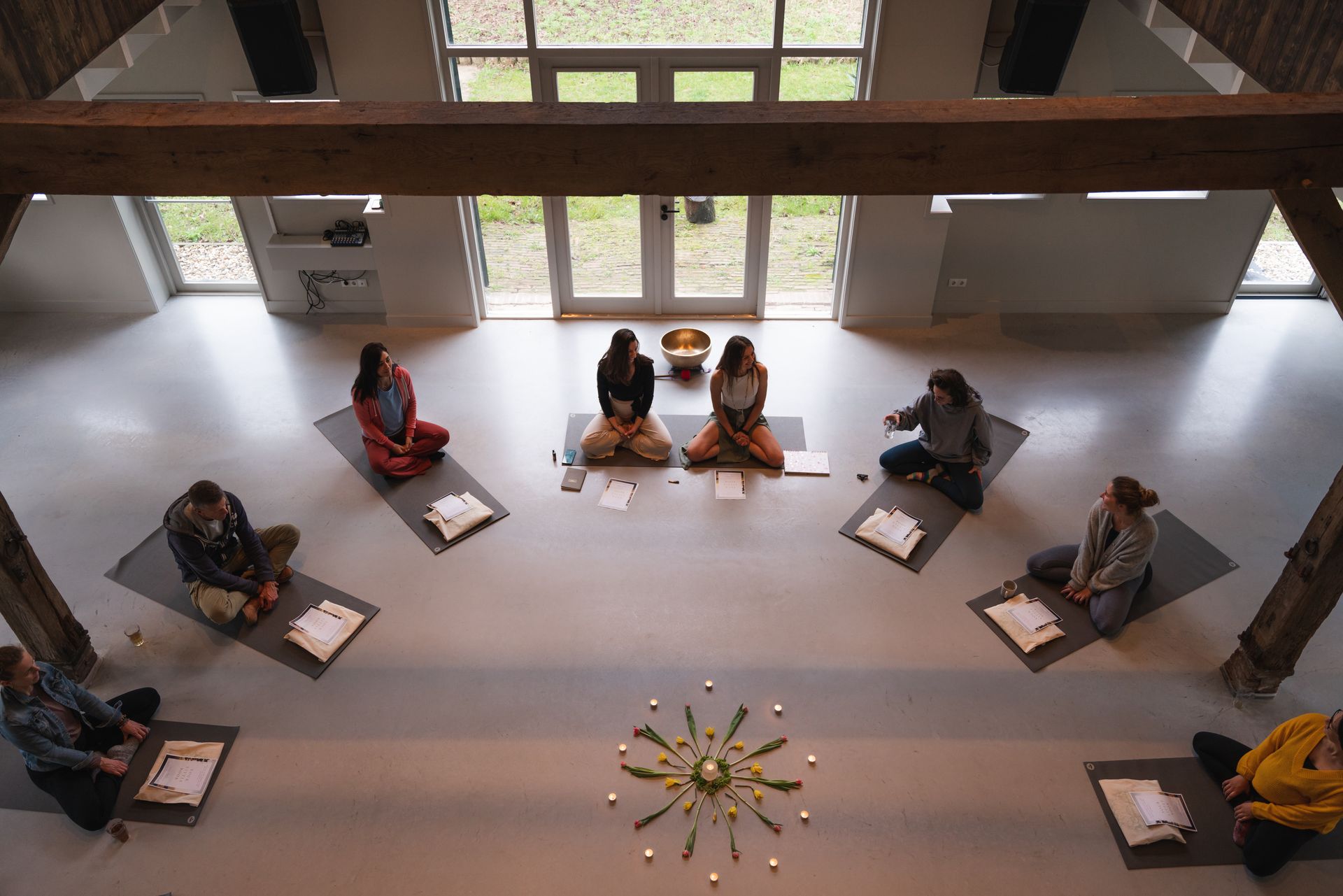 People in a circle meditating on mats, inside a building with large windows and wooden beams.