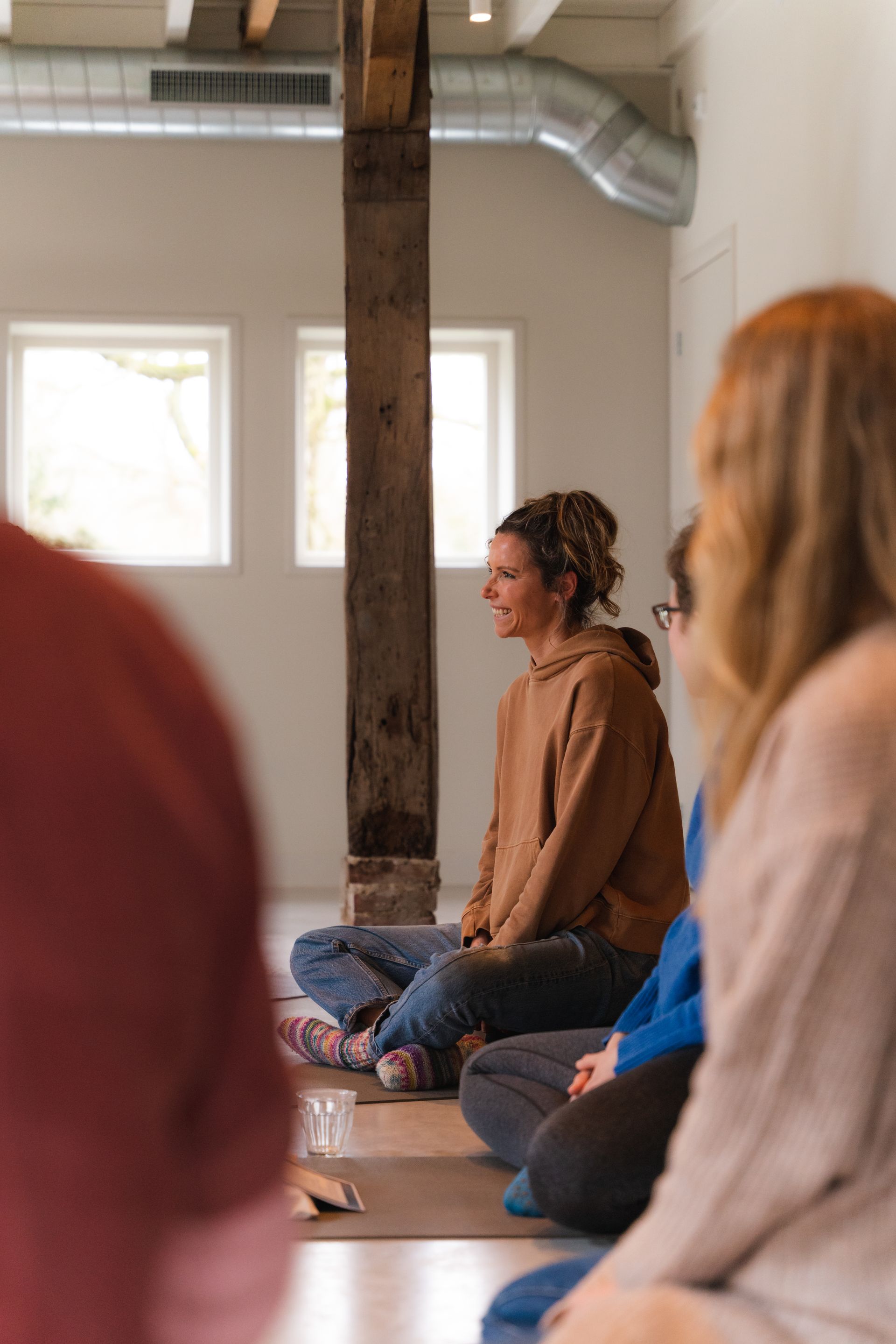 People meditating in a light-filled room; woman in brown sweater sits cross-legged, smiling.