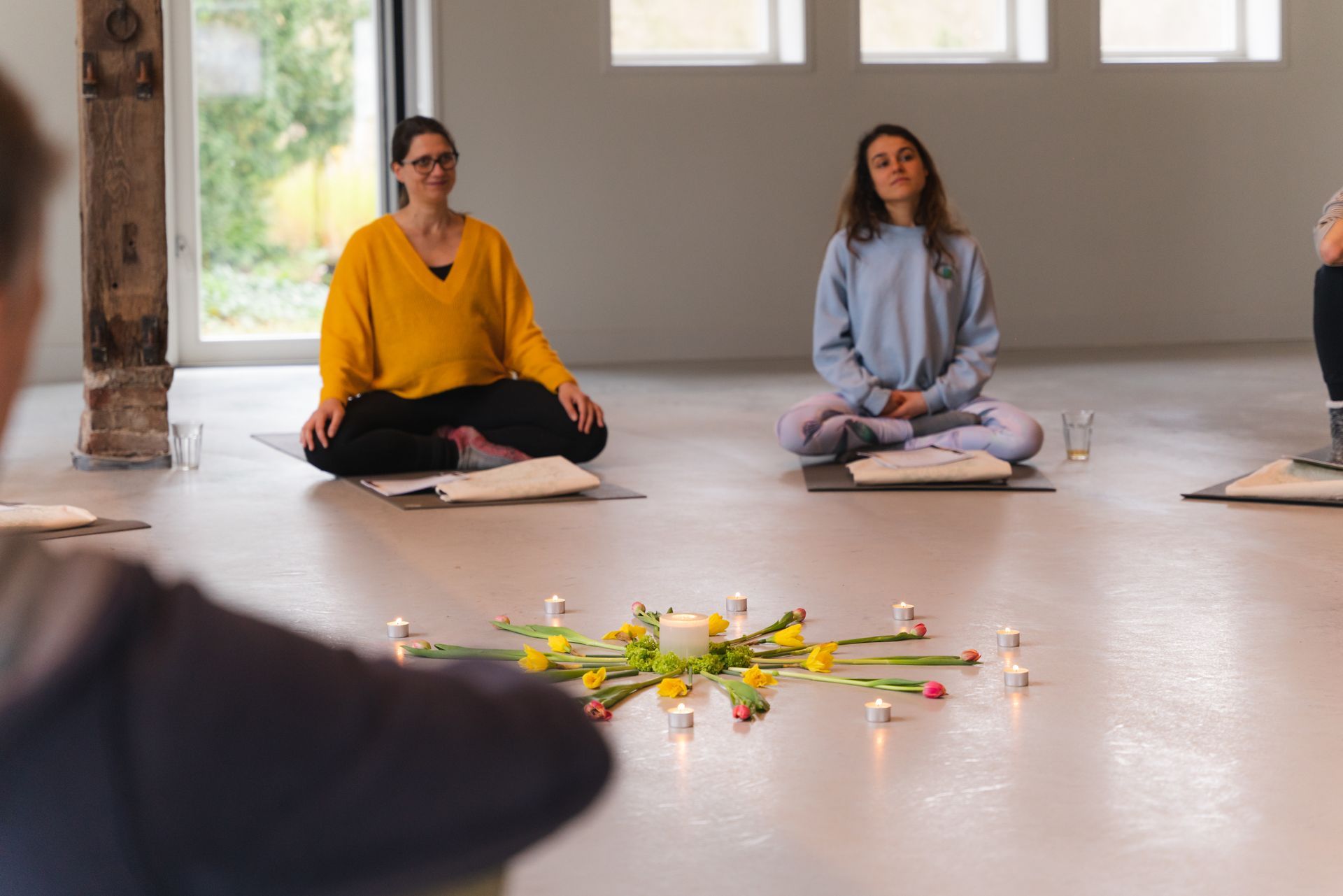 People meditating in a brightly lit room, arranged around a flower and candle mandala.