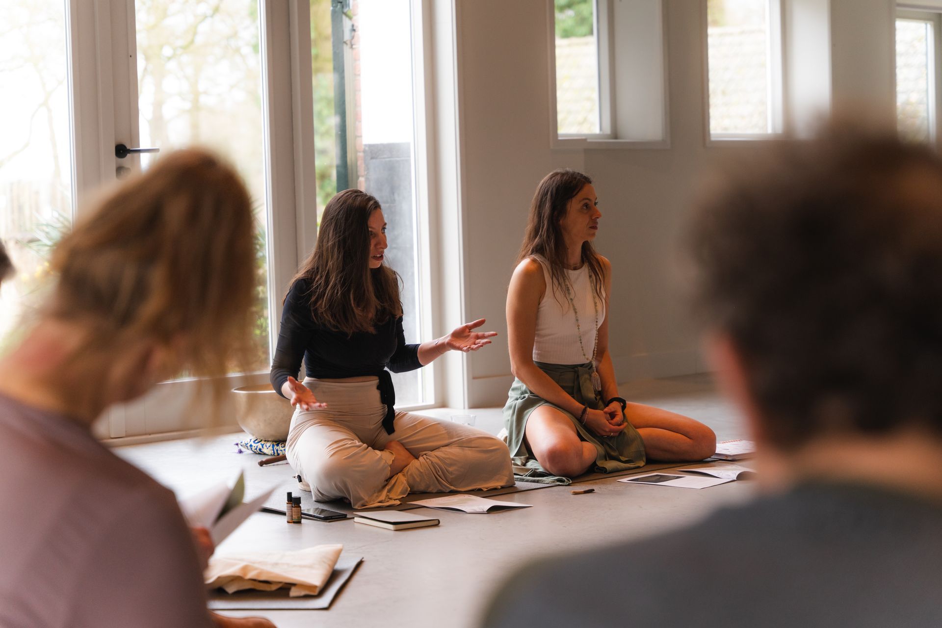 Two women seated on the floor, hands gesturing, leading a group session in a bright room.