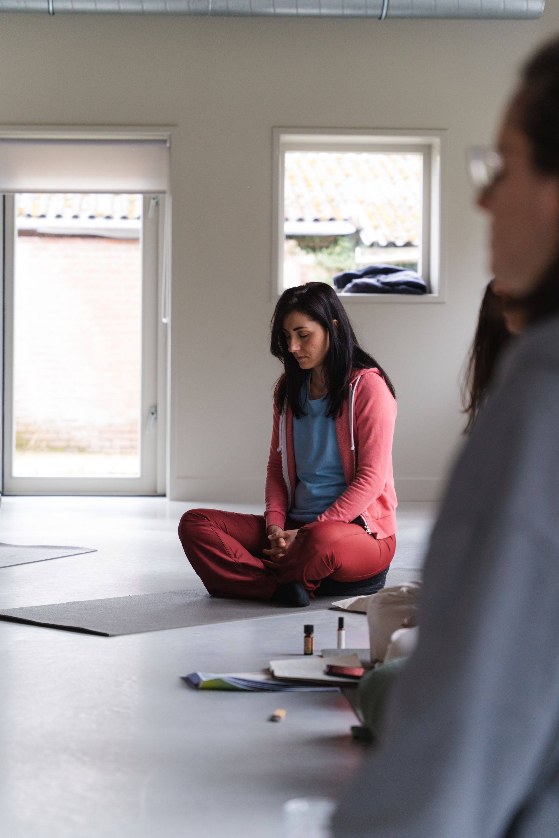Woman meditating indoors, eyes closed, legs crossed, wearing pink jacket and red pants.