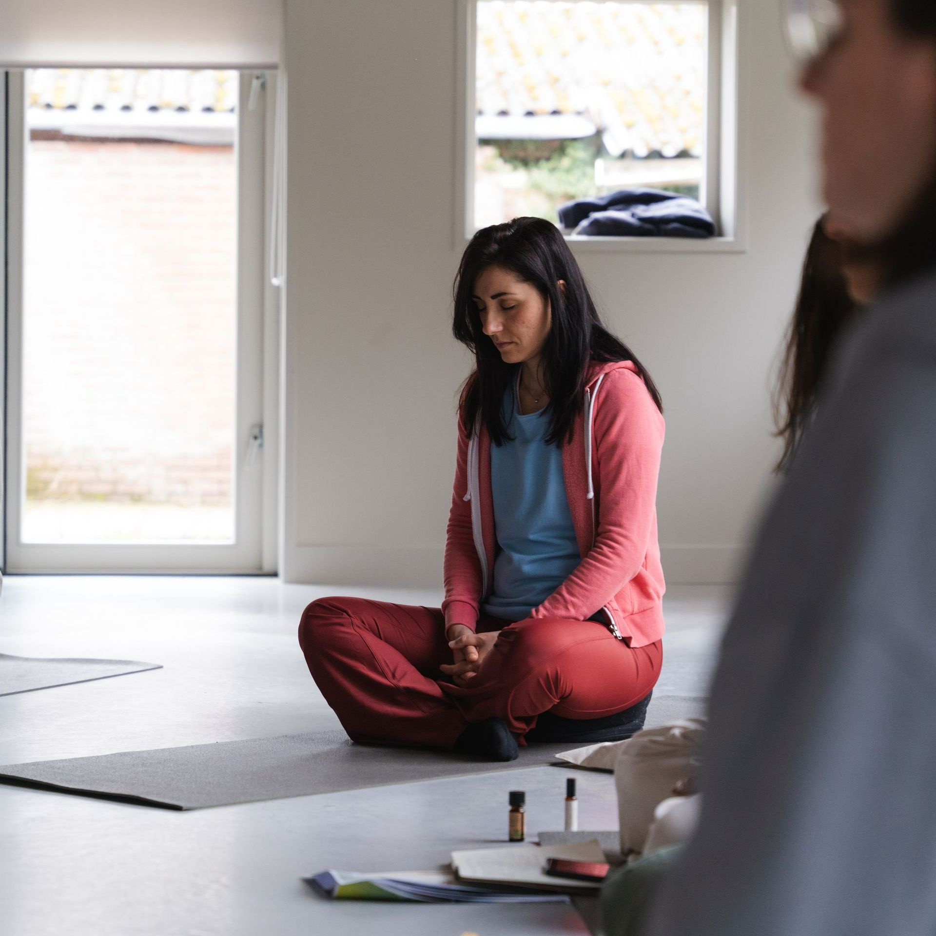 Woman sitting cross-legged on mat, eyes closed. Wearing red pants and a pink jacket, in a bright room.