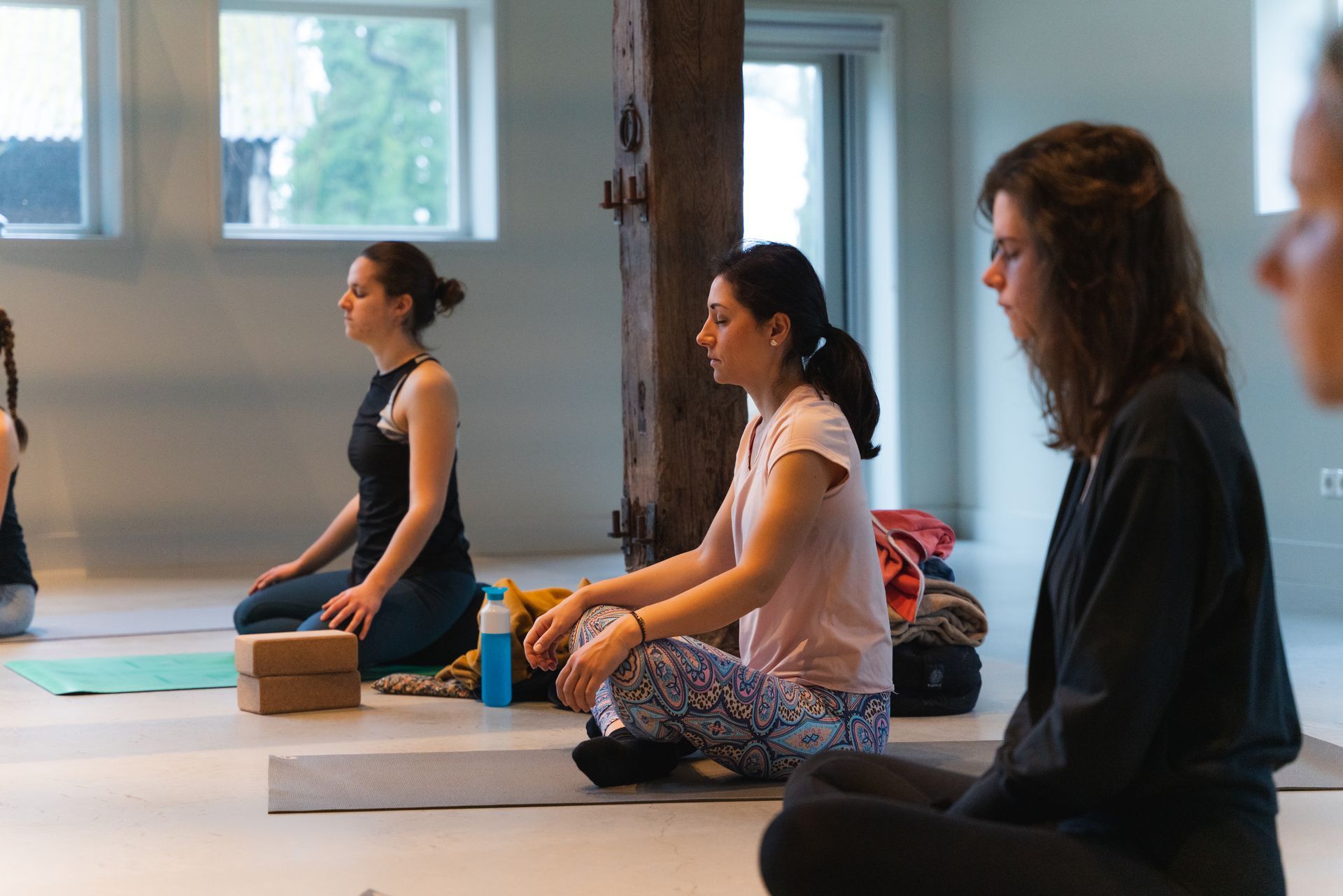 People meditating in a yoga studio, sitting cross-legged with eyes closed.