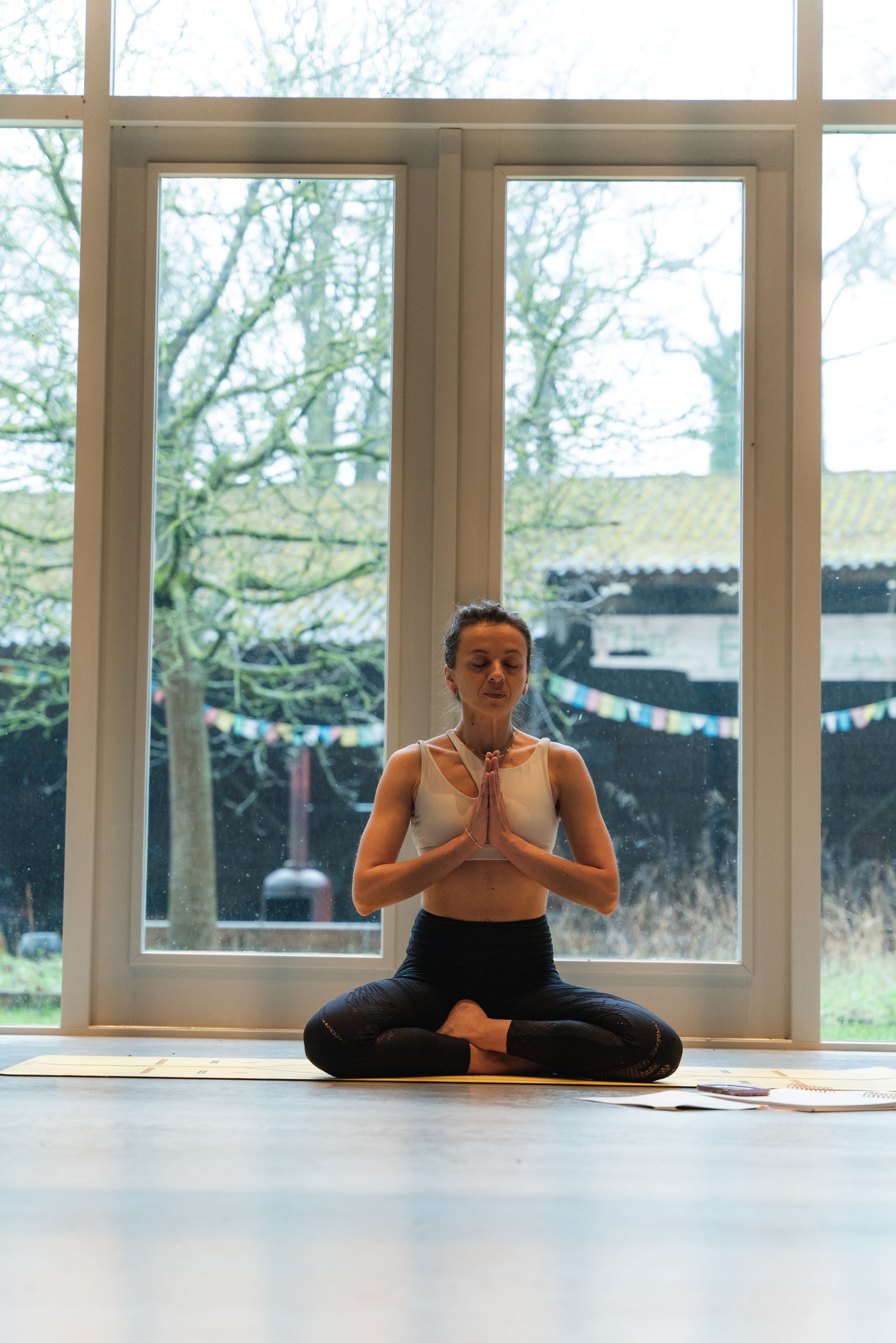 Woman in lotus position, hands in prayer, meditating in front of a window.