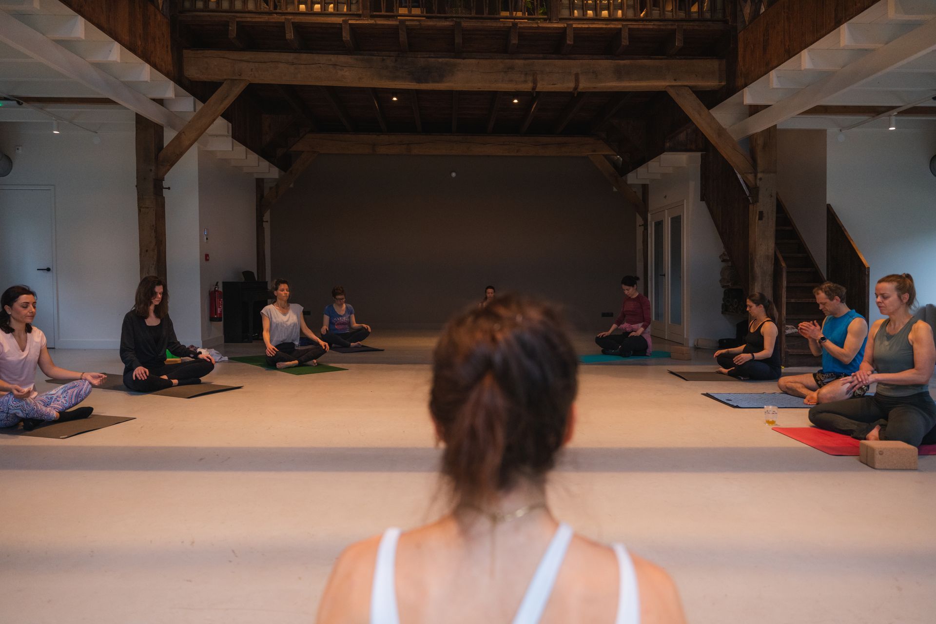 Yoga class: People meditating on mats in a bright studio with wood beams, led by a person in the foreground.
