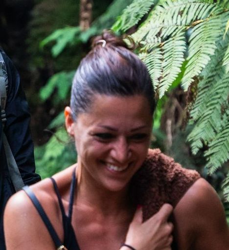 Woman smiling, with dark hair in a bun, wearing black straps. Brown towel on her shoulder. Ferns in the background.