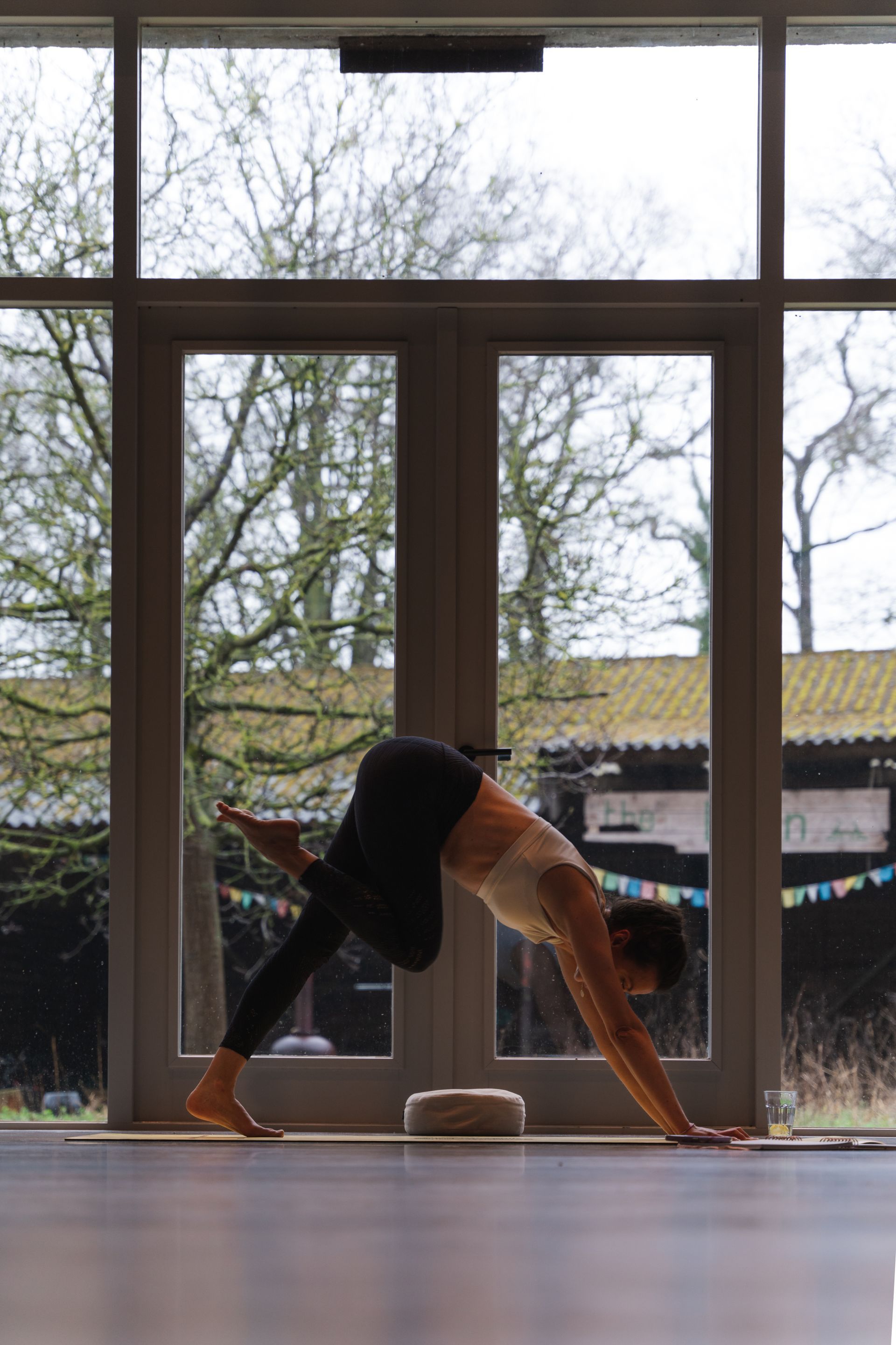 Woman in yoga pose in front of a window, arms extended, leg raised, on a wooden floor.