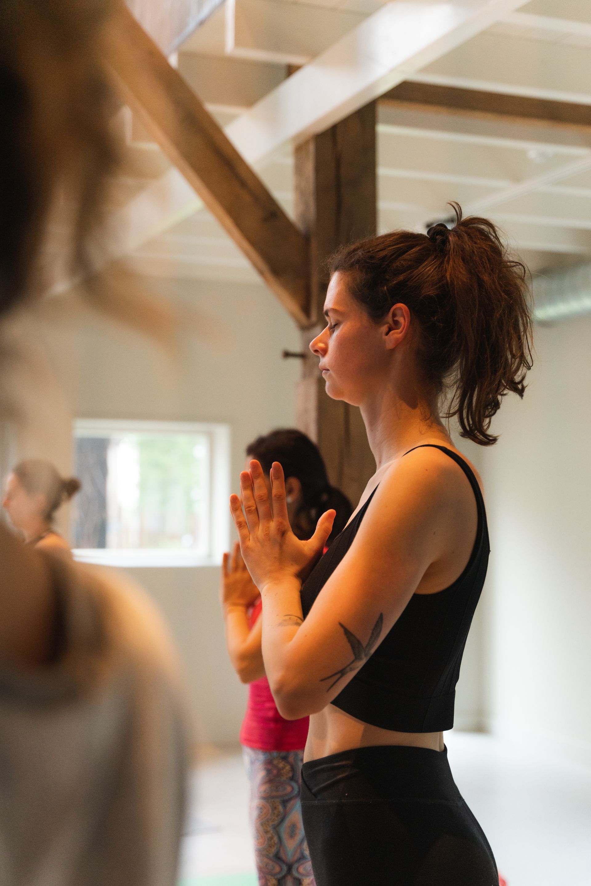 Woman in black yoga outfit meditates with hands in prayer position, indoors with others.