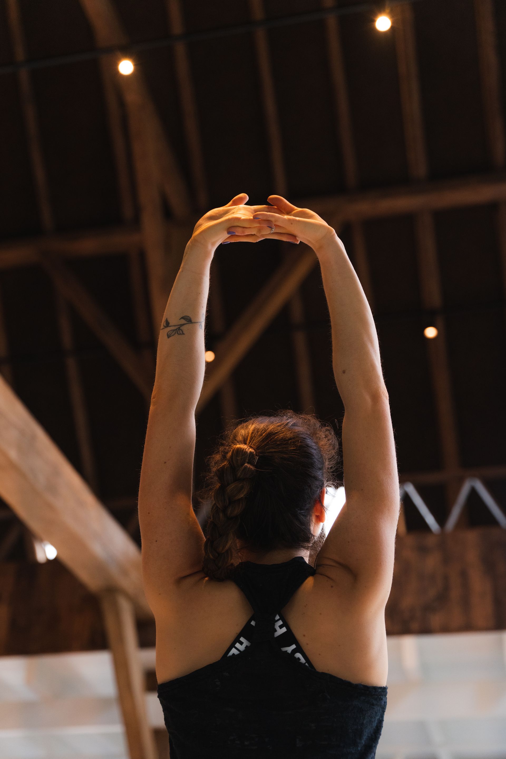 Woman in black tank top stretches arms overhead in a wooden-beamed structure.