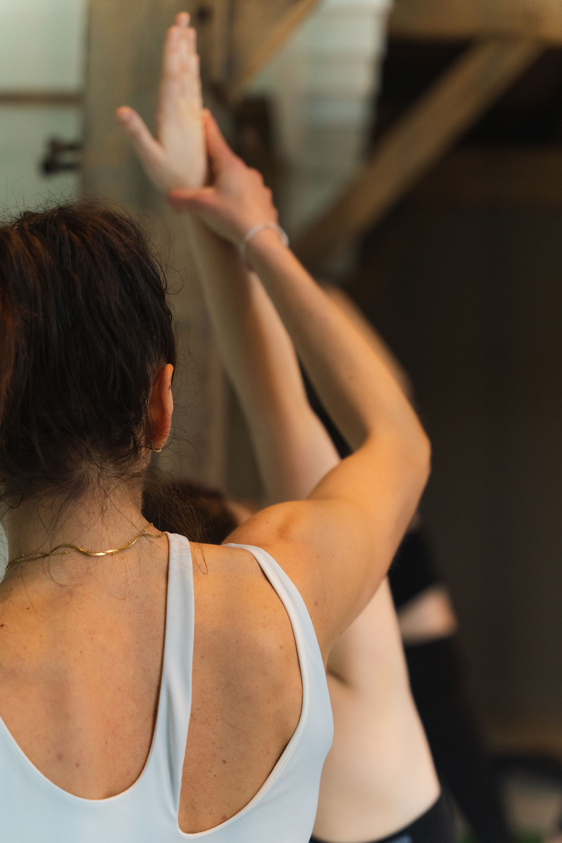 Woman assisting another in yoga, reaching arms up in a sunlit room.
