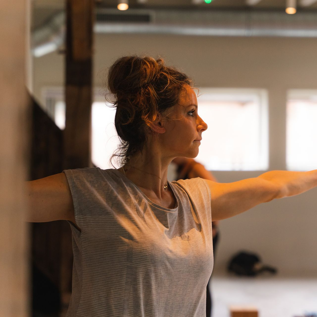 Woman in a gray shirt doing yoga with arms outstretched in a bright studio.