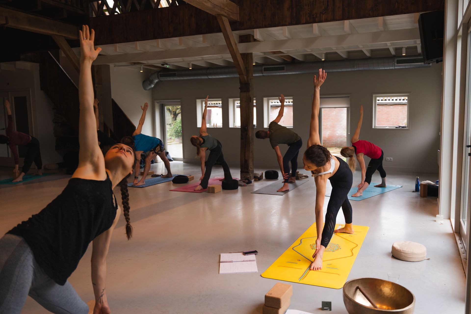 People practice yoga in a studio with high ceilings. Several people in athletic wear are in the Triangle Pose.