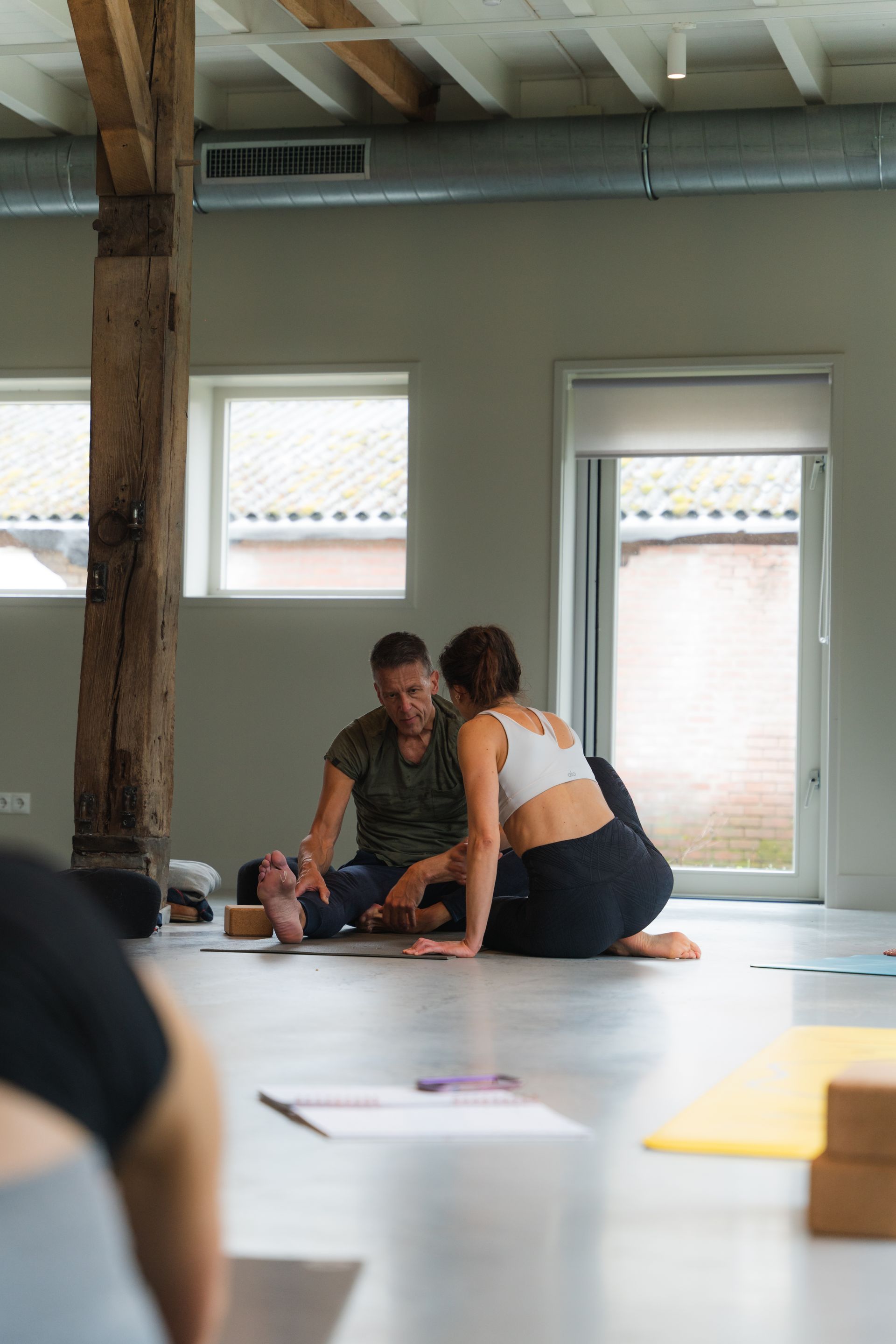 Yoga instructor assisting student on the floor, in a bright studio with wood beams.