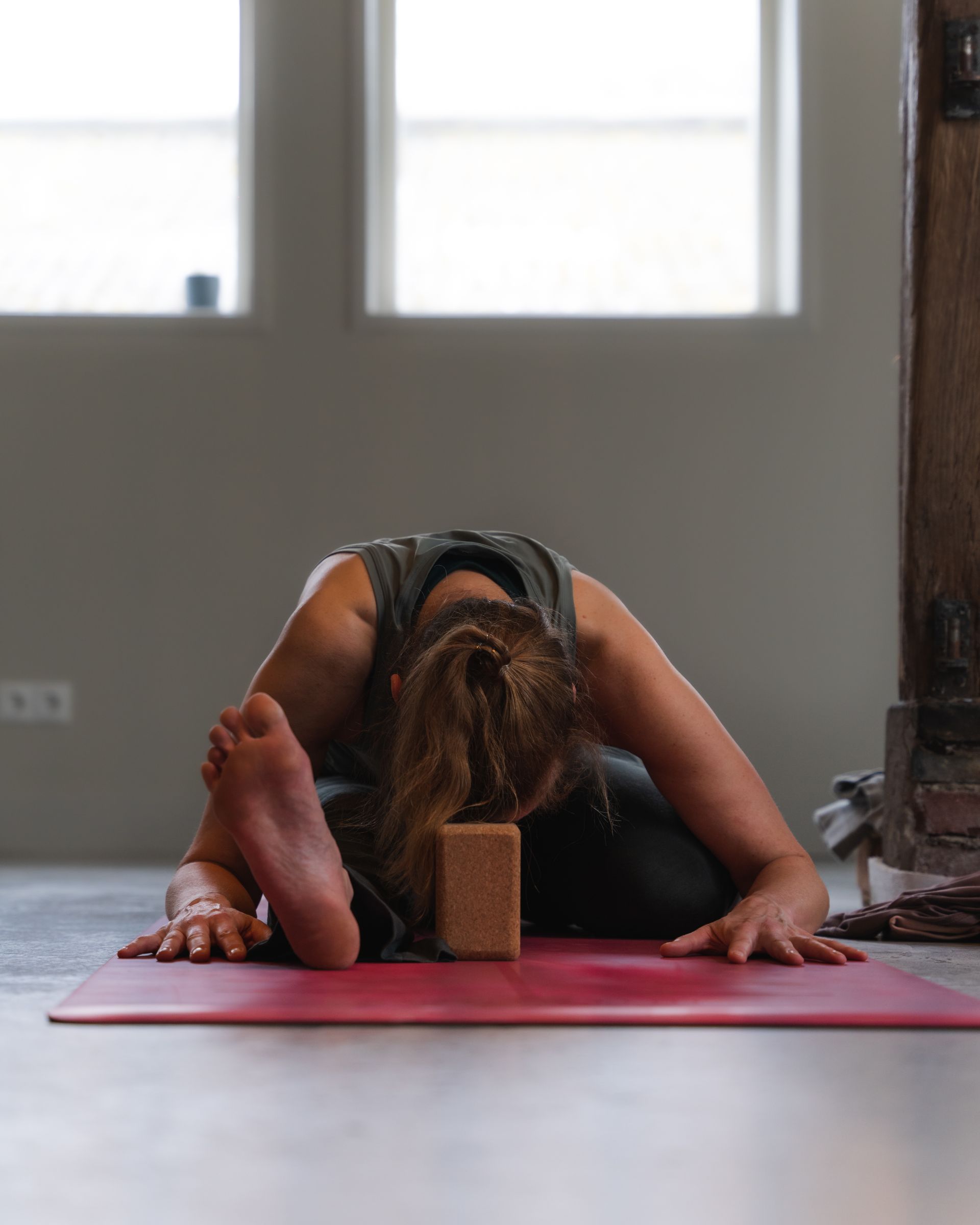 Person in seated yoga pose, leaning forward with foot extended, hands on mat, wooden block in front.