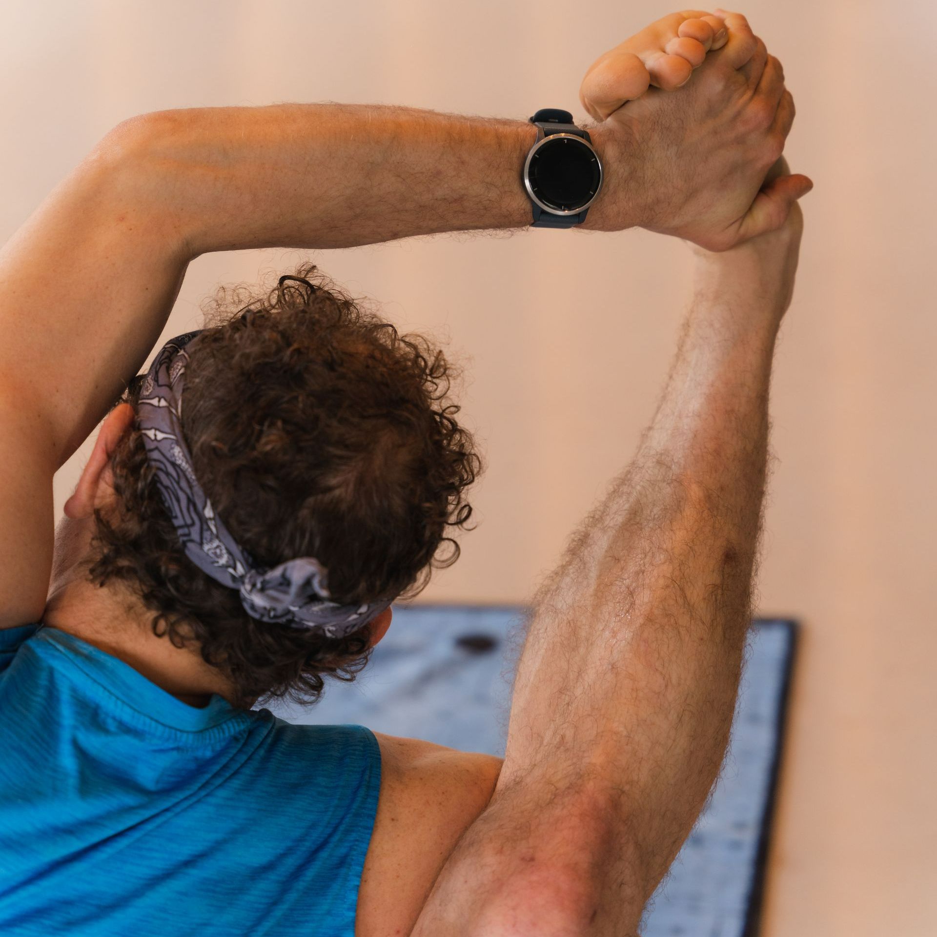 Person stretching, arms overhead, wearing a watch and a bandana. Blue shirt, indoors.