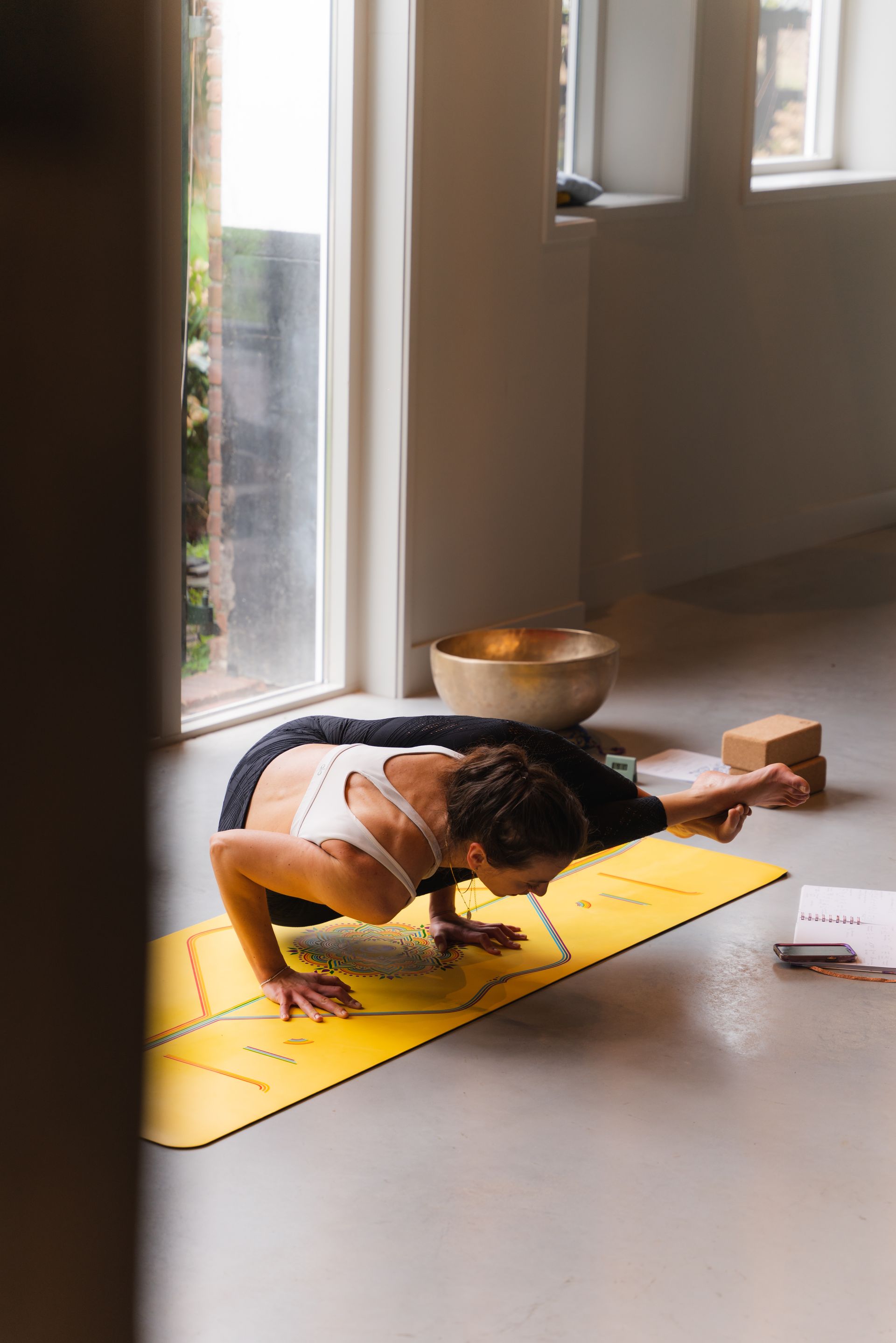 Woman in a yoga pose on a yellow mat, extending one arm, in a bright room near a window.