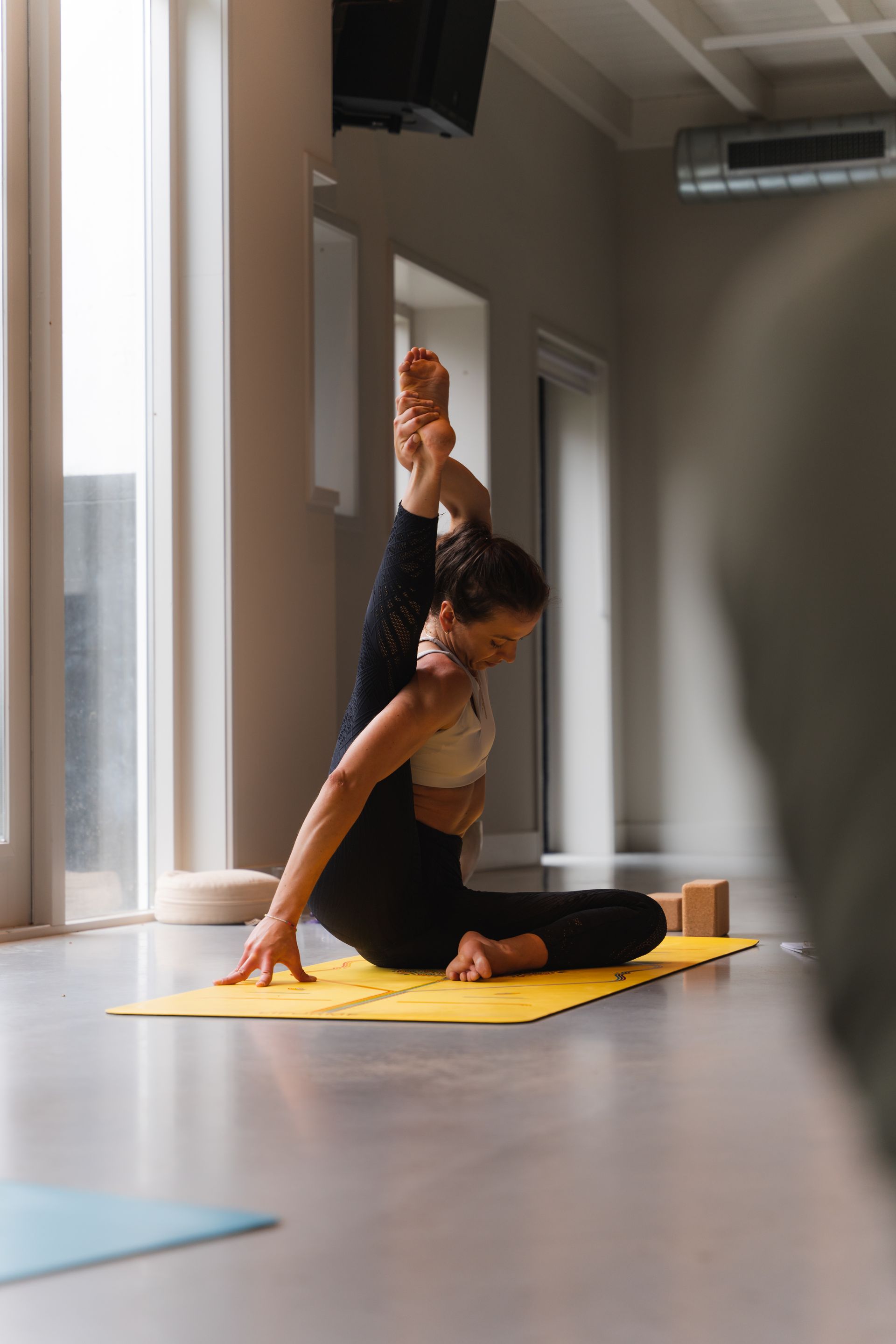 Woman doing yoga with leg extended overhead on a yellow mat. Neutral indoor setting.