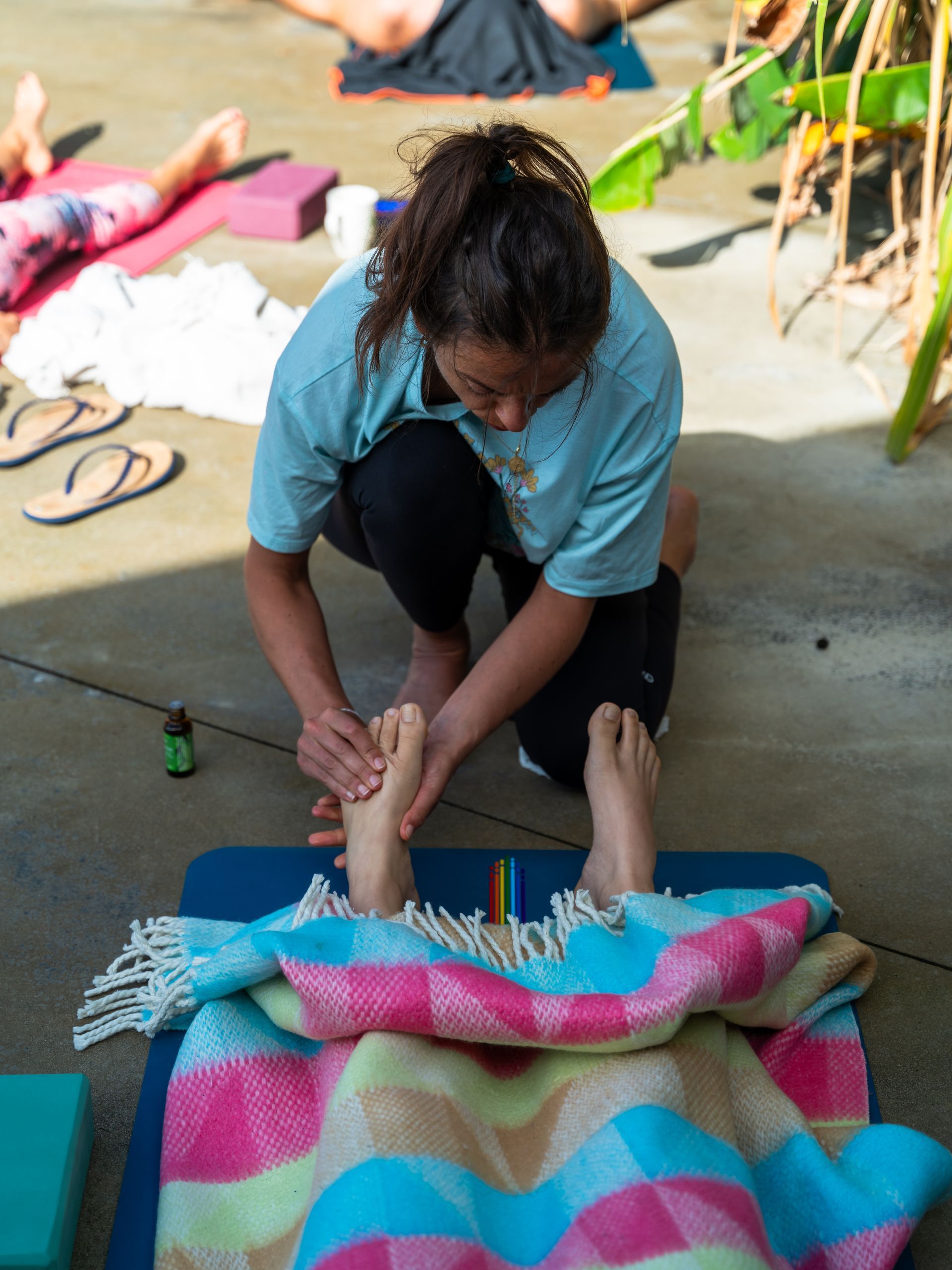 Person massaging someone's feet covered by a colorful blanket during outdoor yoga.