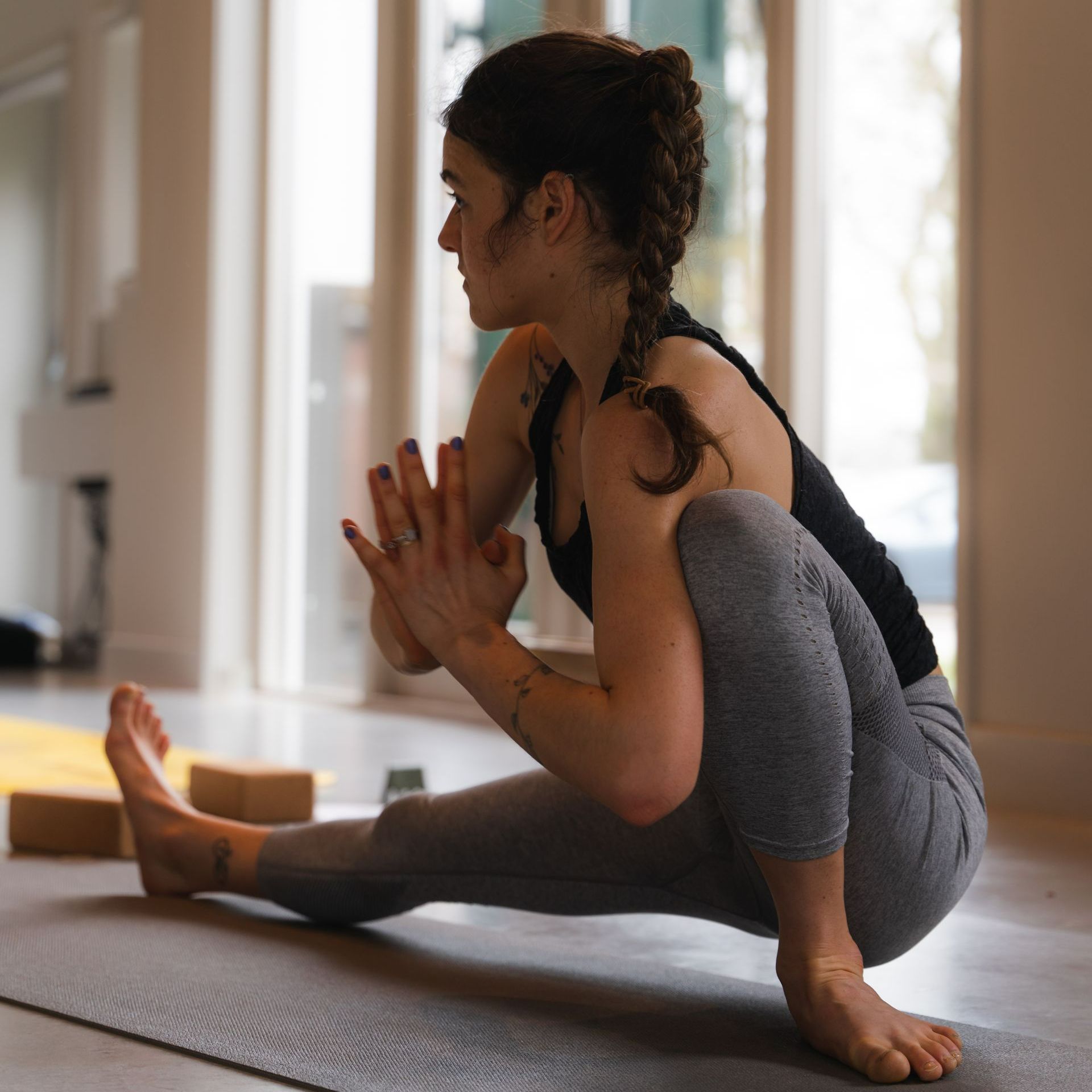 Woman in yoga pose, hands in prayer position, on a mat in a light-filled room.