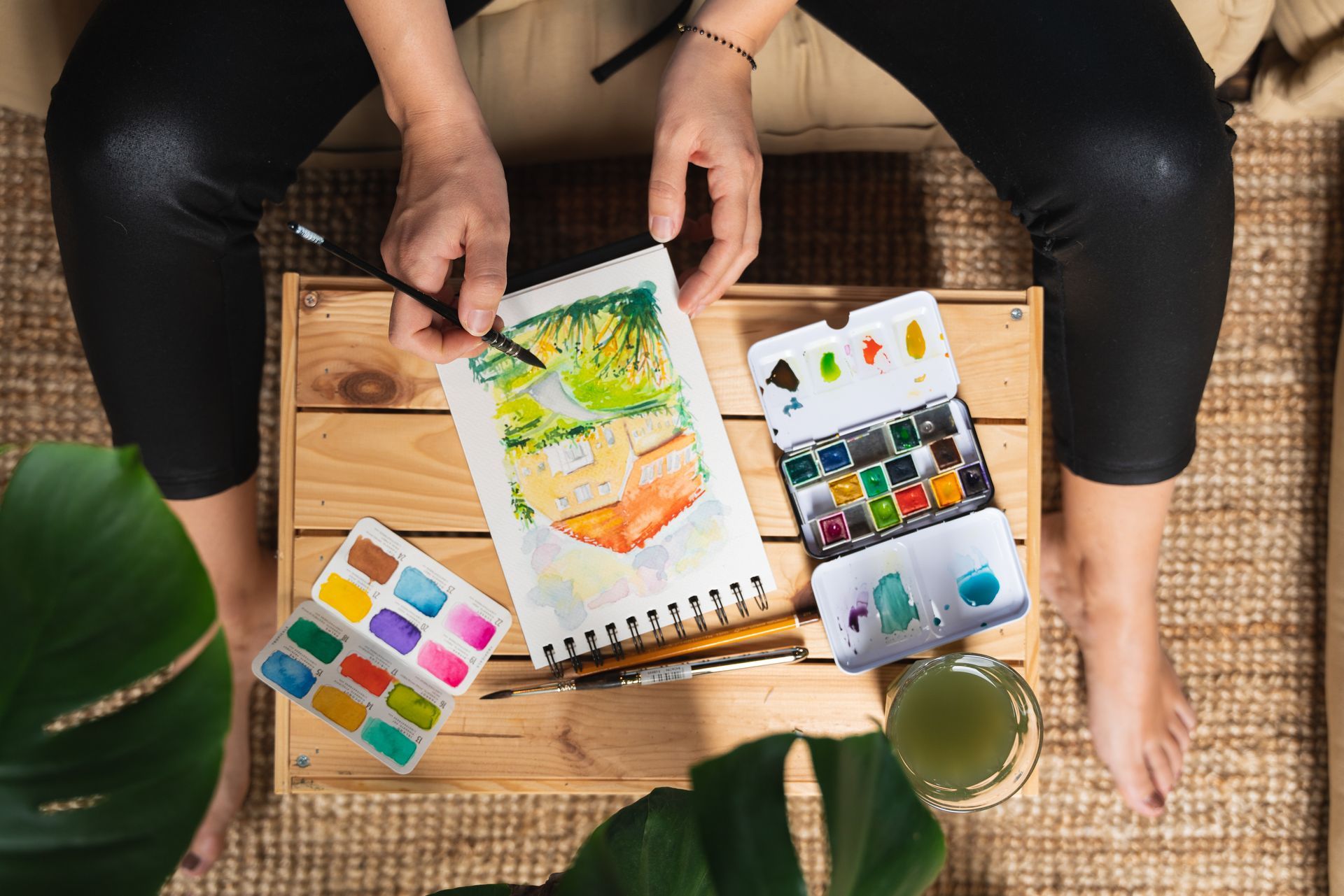 Person painting with watercolors on a wooden crate, surrounded by art supplies and a plant.