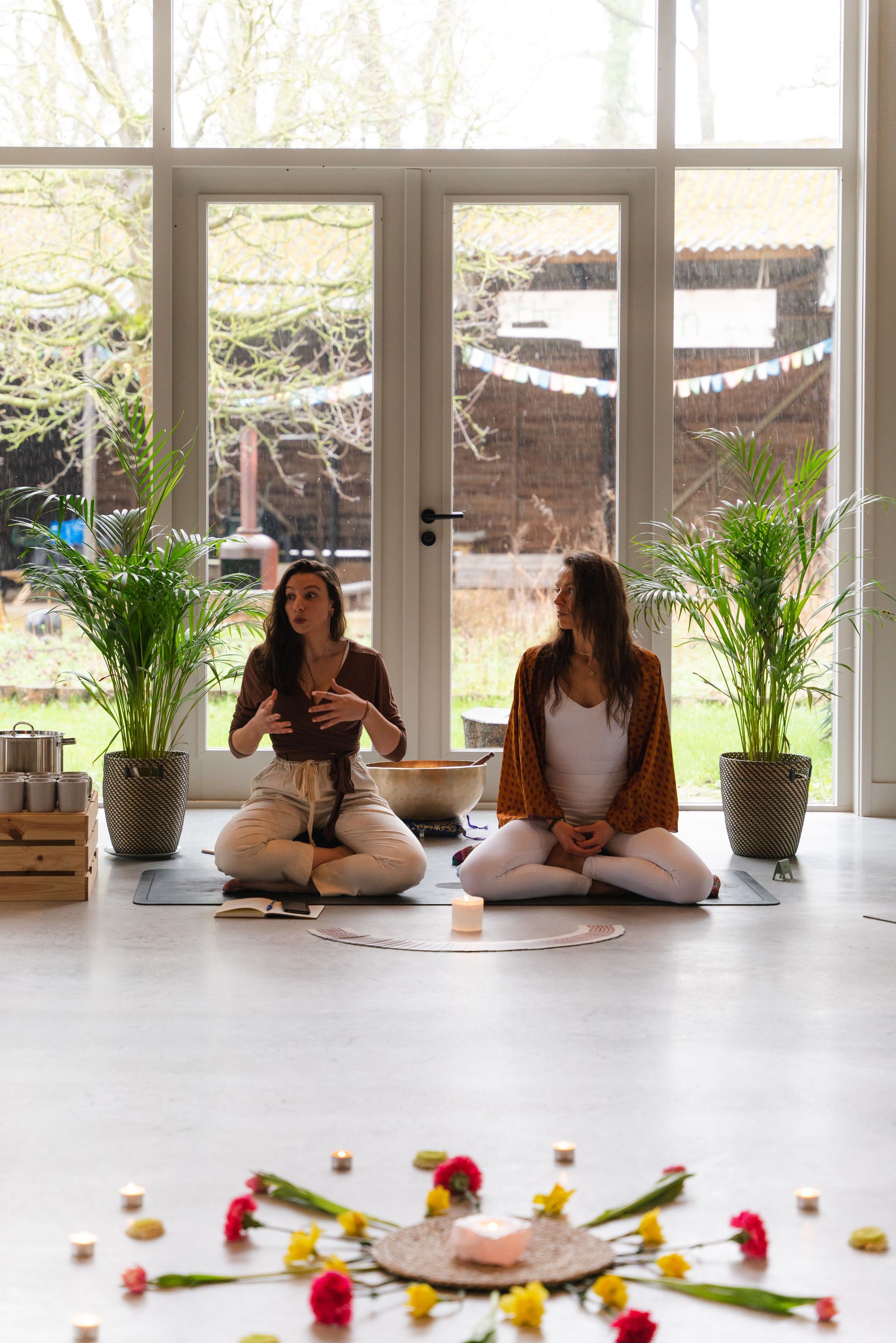 Two people in lotus position, inside a bright room, surrounded by flowers and candles.