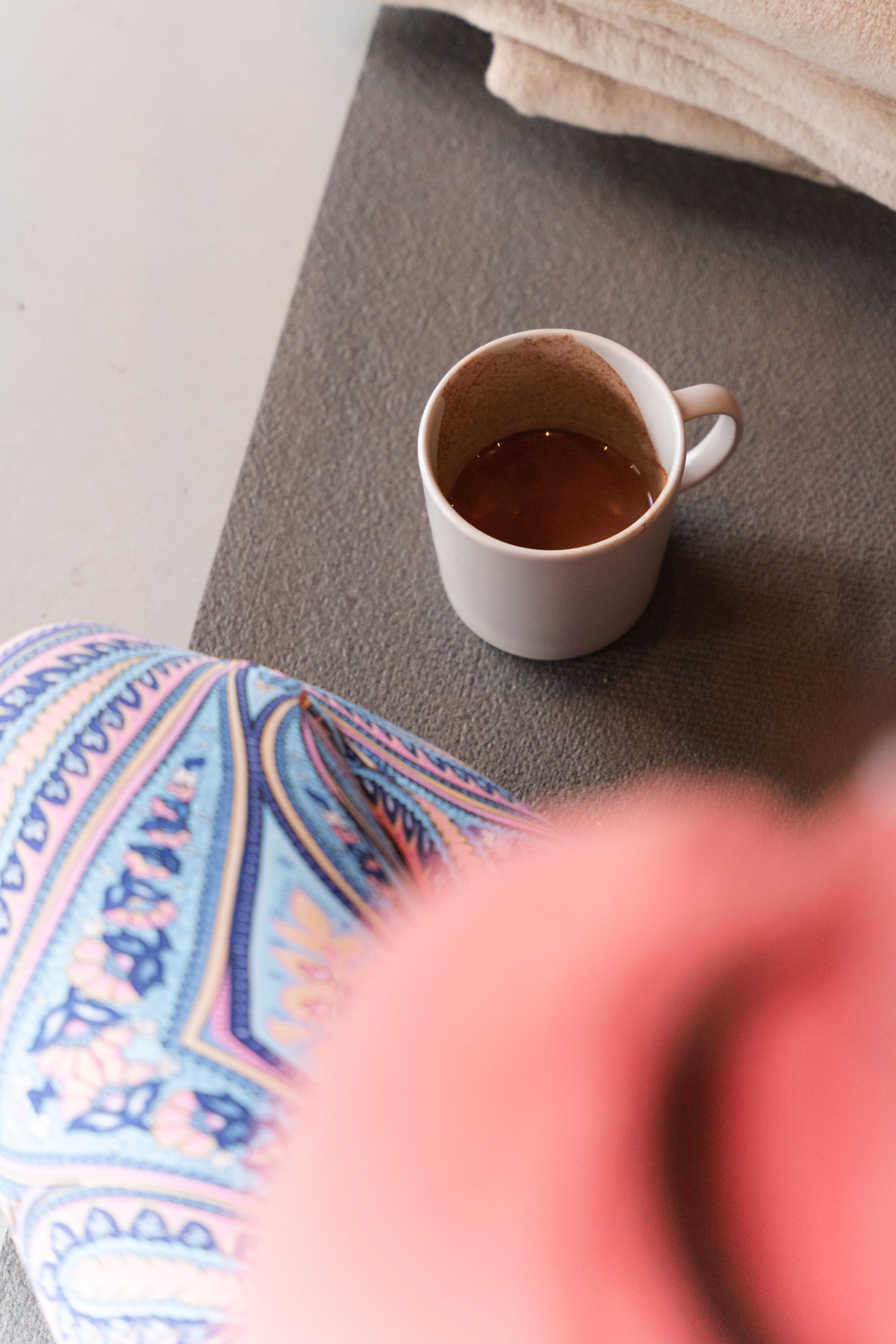 White mug with brown liquid on a gray surface, patterned pants in the foreground, and a peach-colored top.