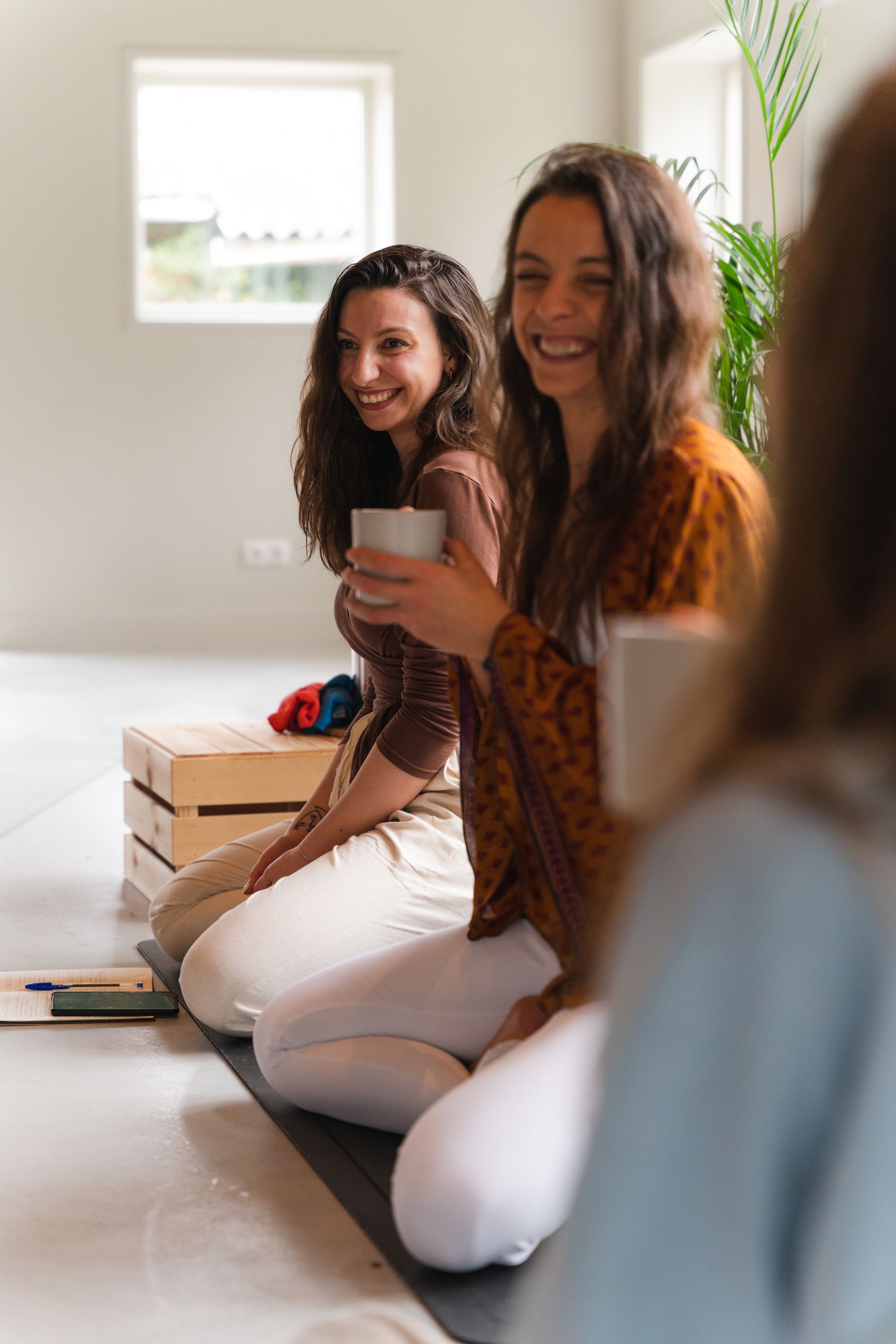 Two smiling women, holding mugs, sit on a mat in a bright room.