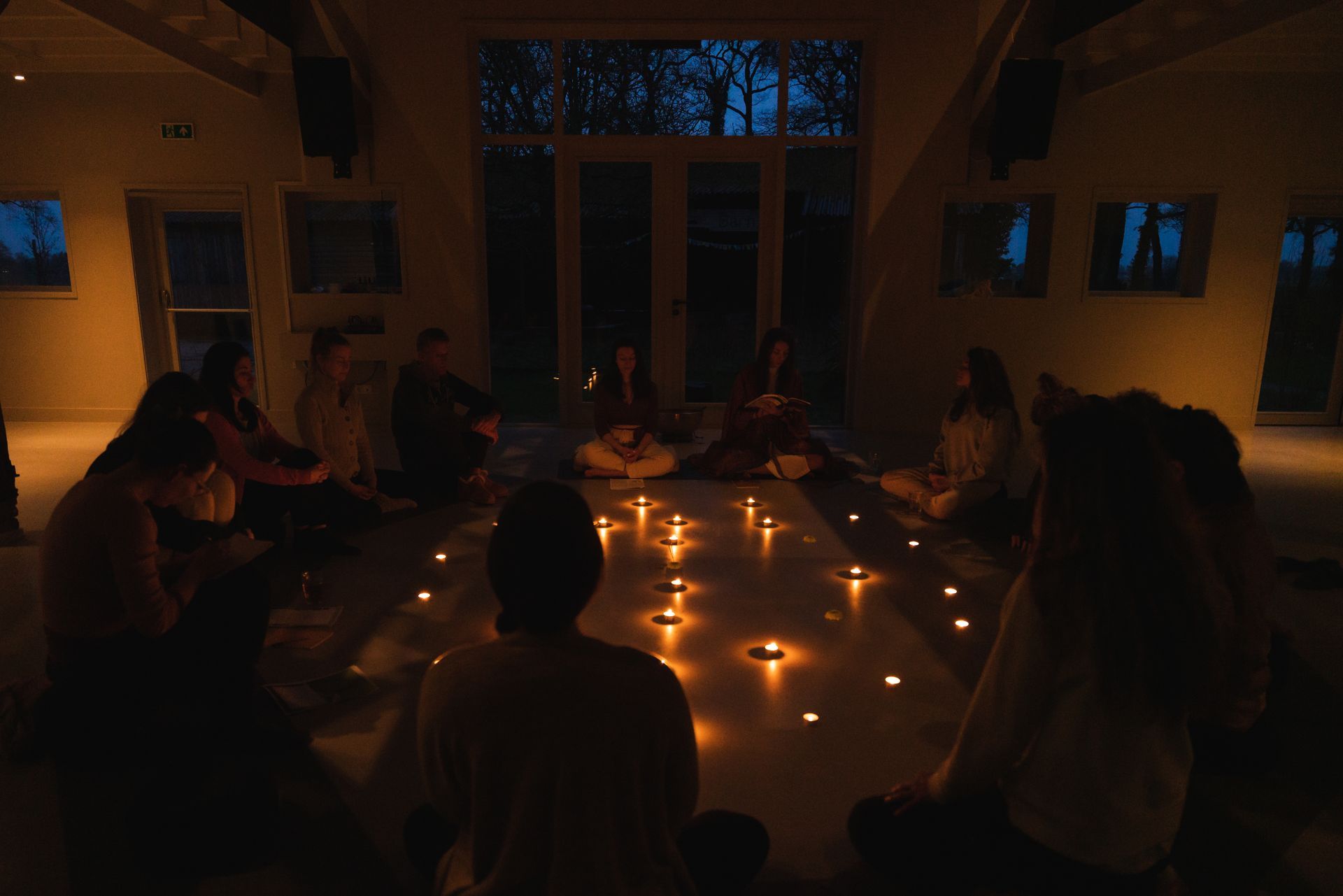 People seated in a circle, meditating by candlelight. Dimly lit room, dark outside windows.