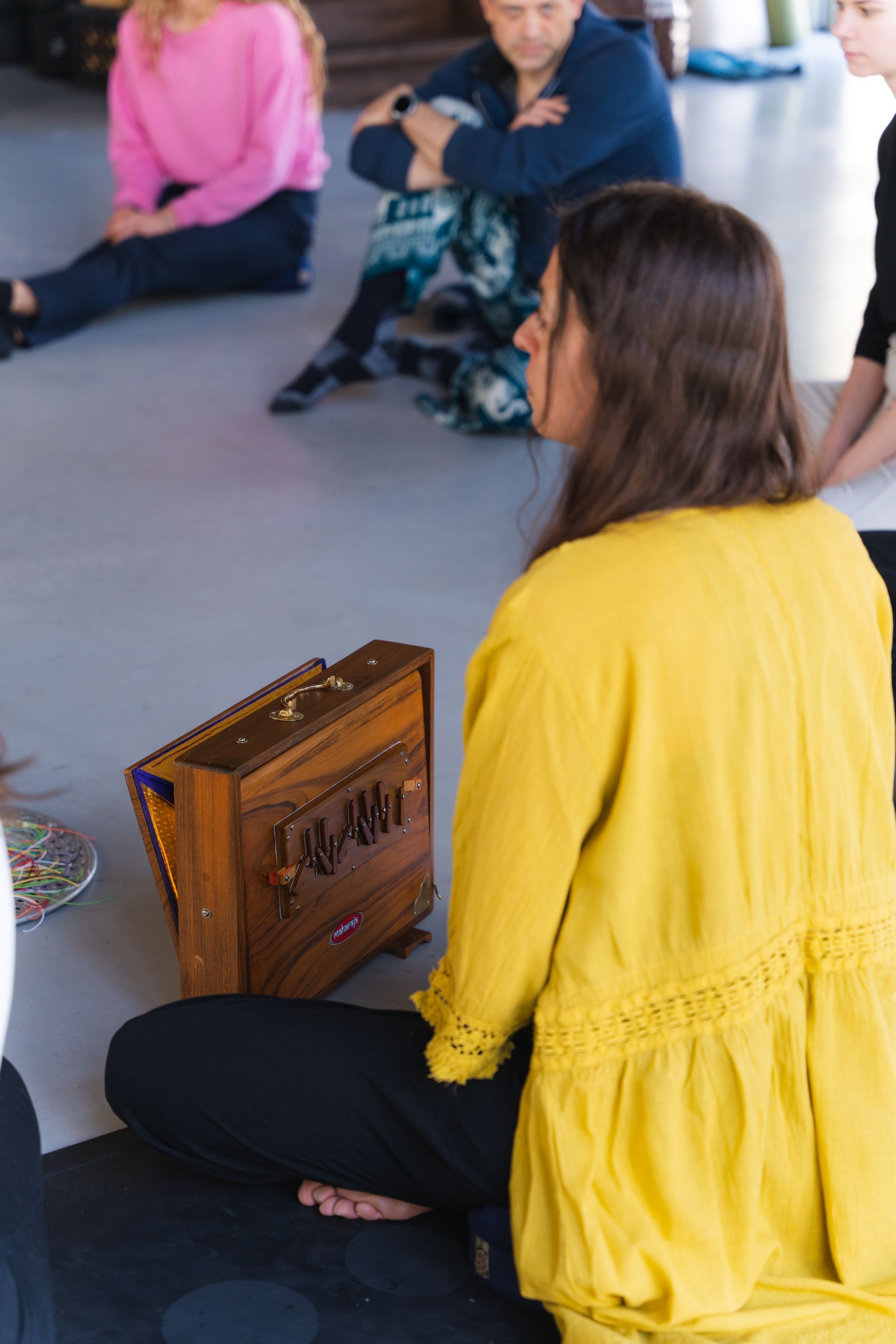 Woman in yellow top sits cross-legged near wooden box in a circle. Others sit on floor.