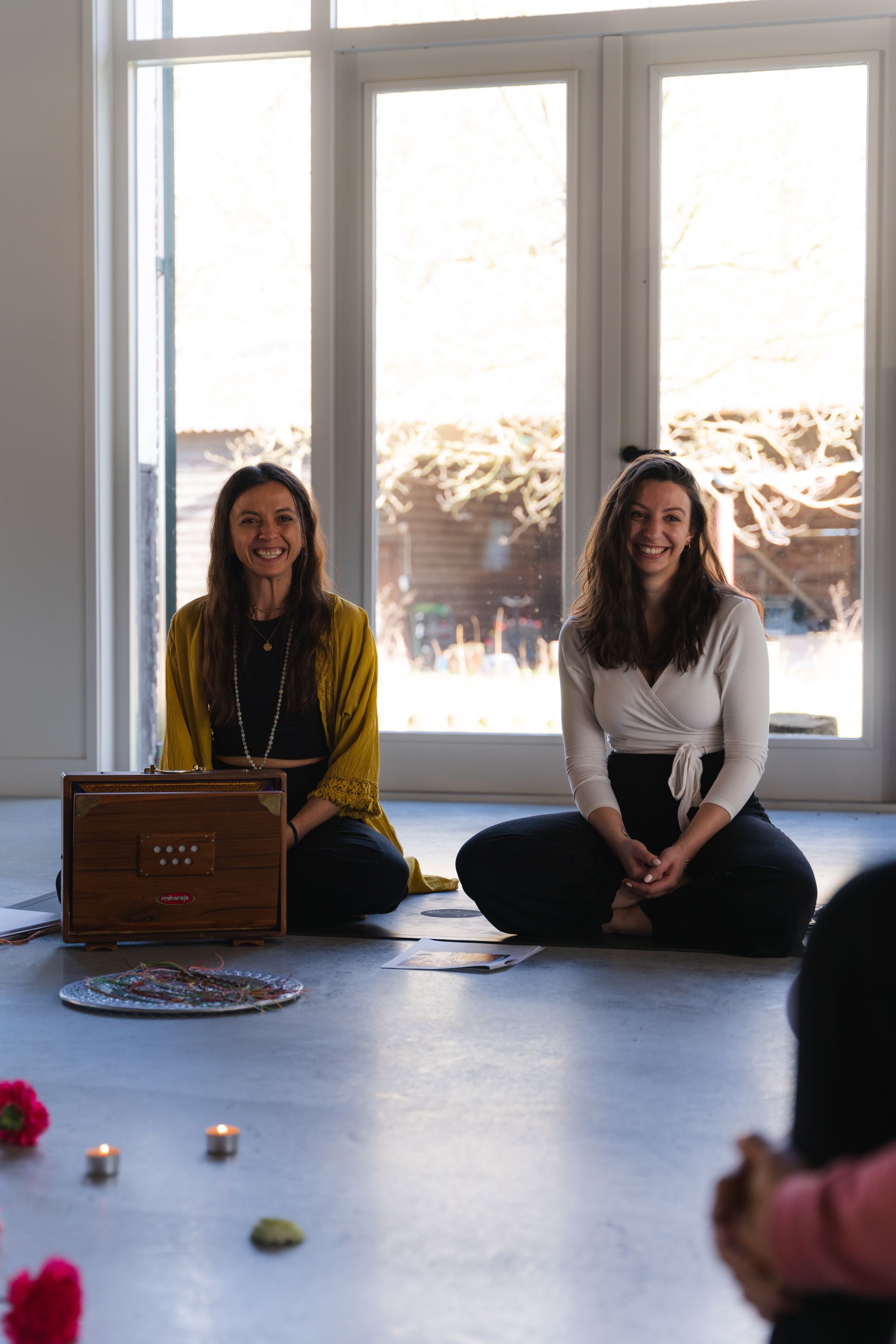 Two smiling women sit on the floor, facing forward, in front of a window. A wooden box is between them.