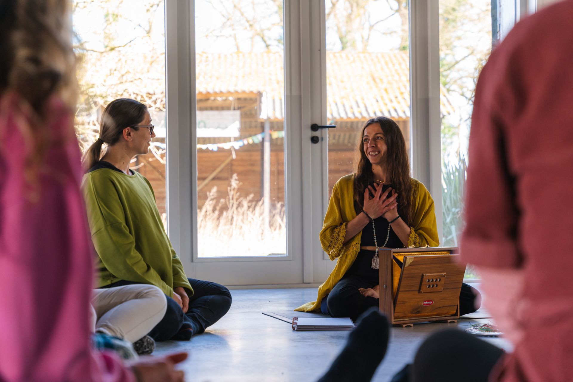 People in circle, meditating in sunlit room; one woman smiles with hands on chest.