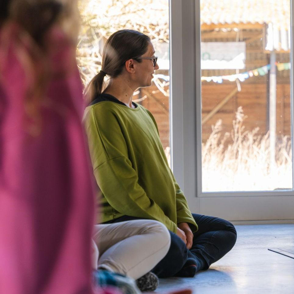 Woman sitting cross-legged indoors, looking out the window. She wears a green sweatshirt, glasses, and has a ponytail.
