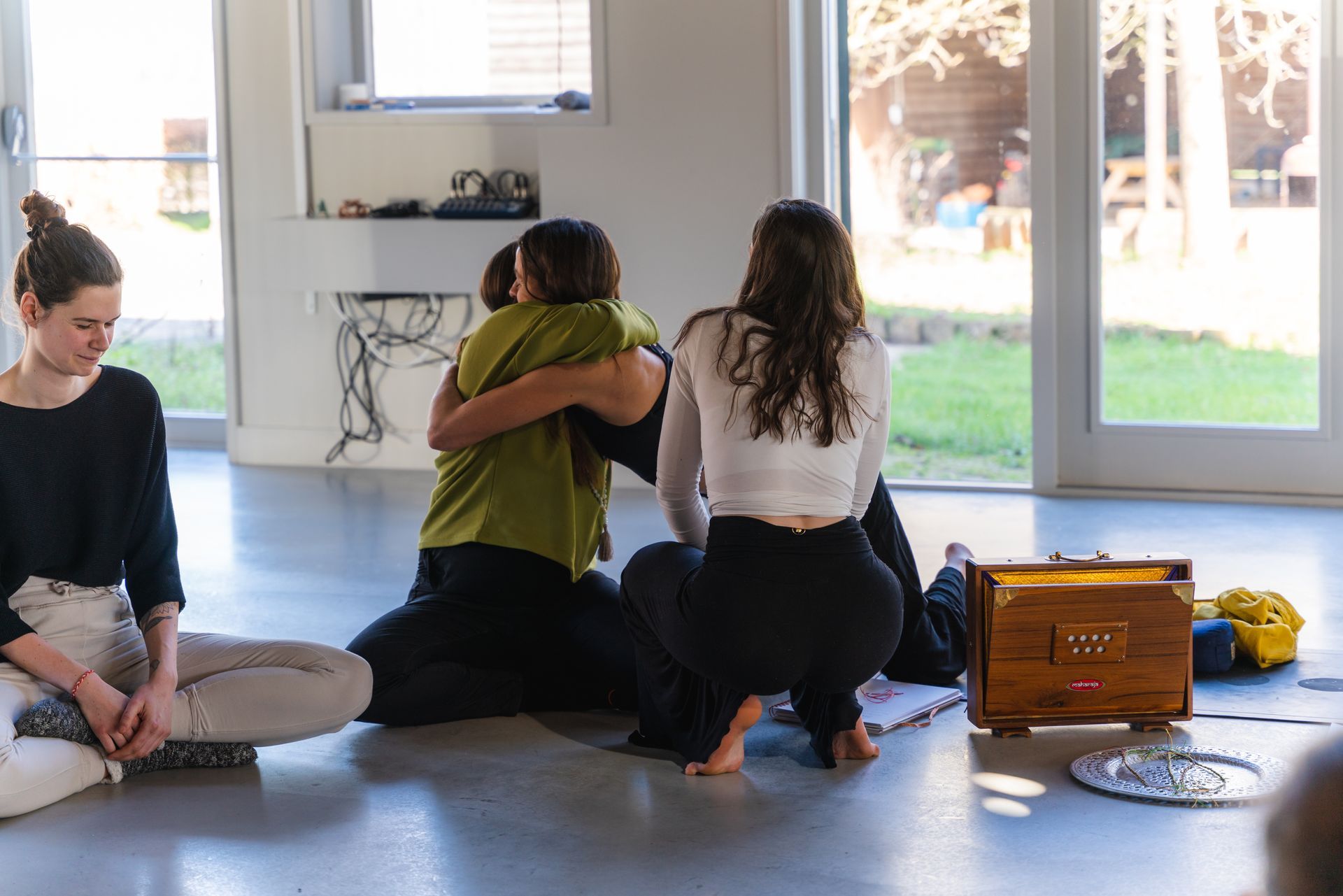 People hugging on the floor of a sunlit room. Two sit near a harmonium and tray.