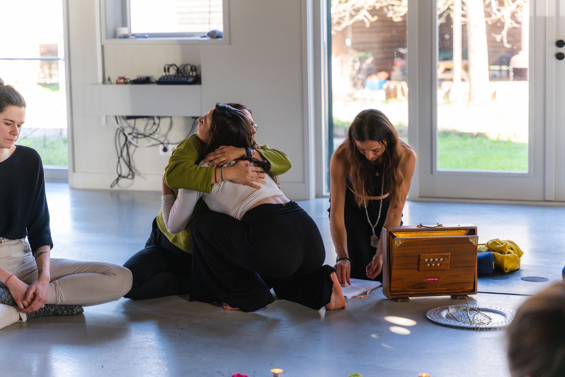People in a circle; two hugging on the floor, one kneeling by a harmonium, others seated. Interior with natural light.